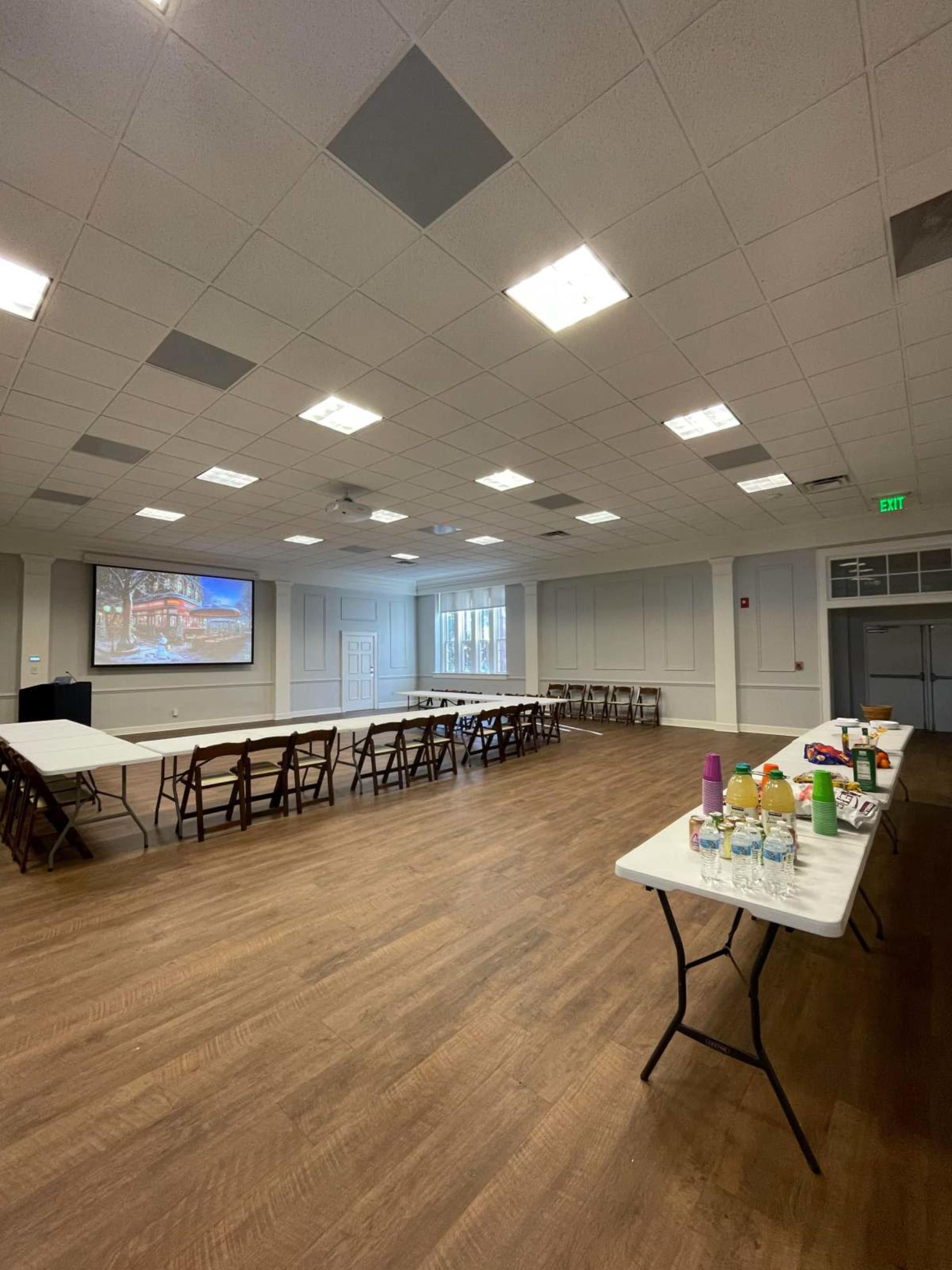 A spacious indoor room features rows of tables set up for an event, with a projector screen on one wall and bright overhead lights illuminating the space.