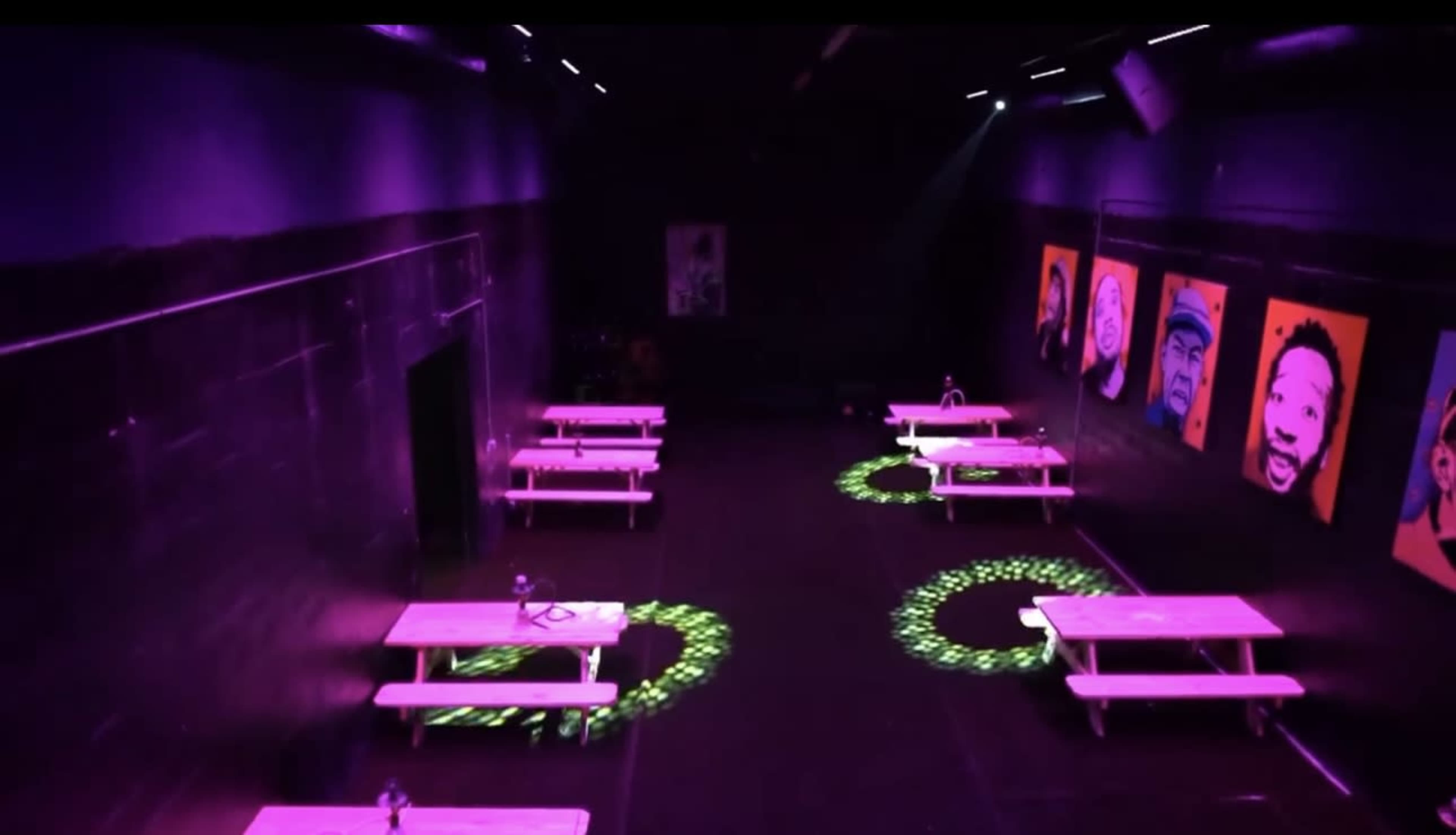 The image shows a dimly lit room with purple lighting, several tables arranged in rows, and colorful portraits hanging on the walls.
