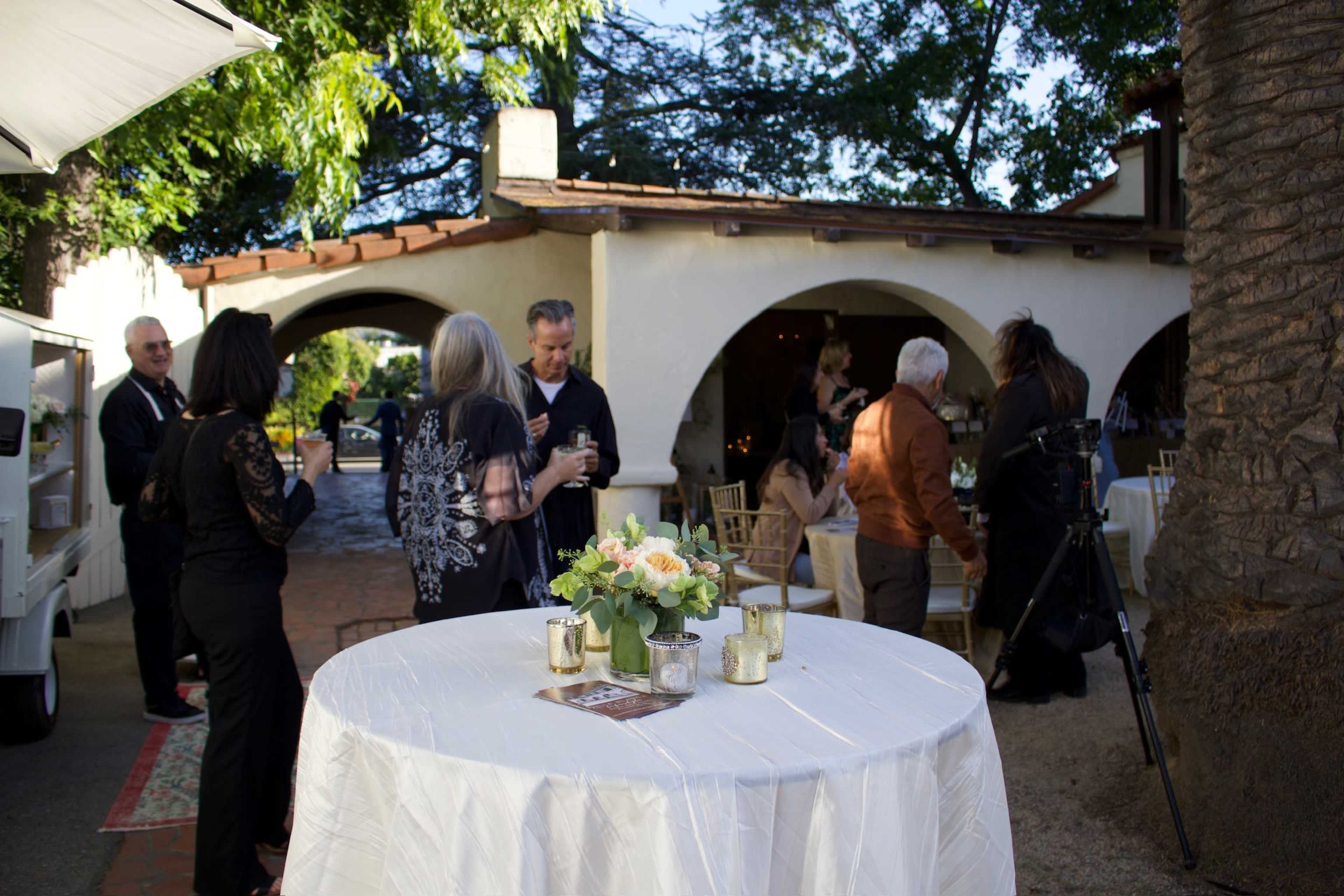 The scene depicts guests mingling at an outdoor event, with elegantly set tables and a backdrop of rustic architecture and greenery.