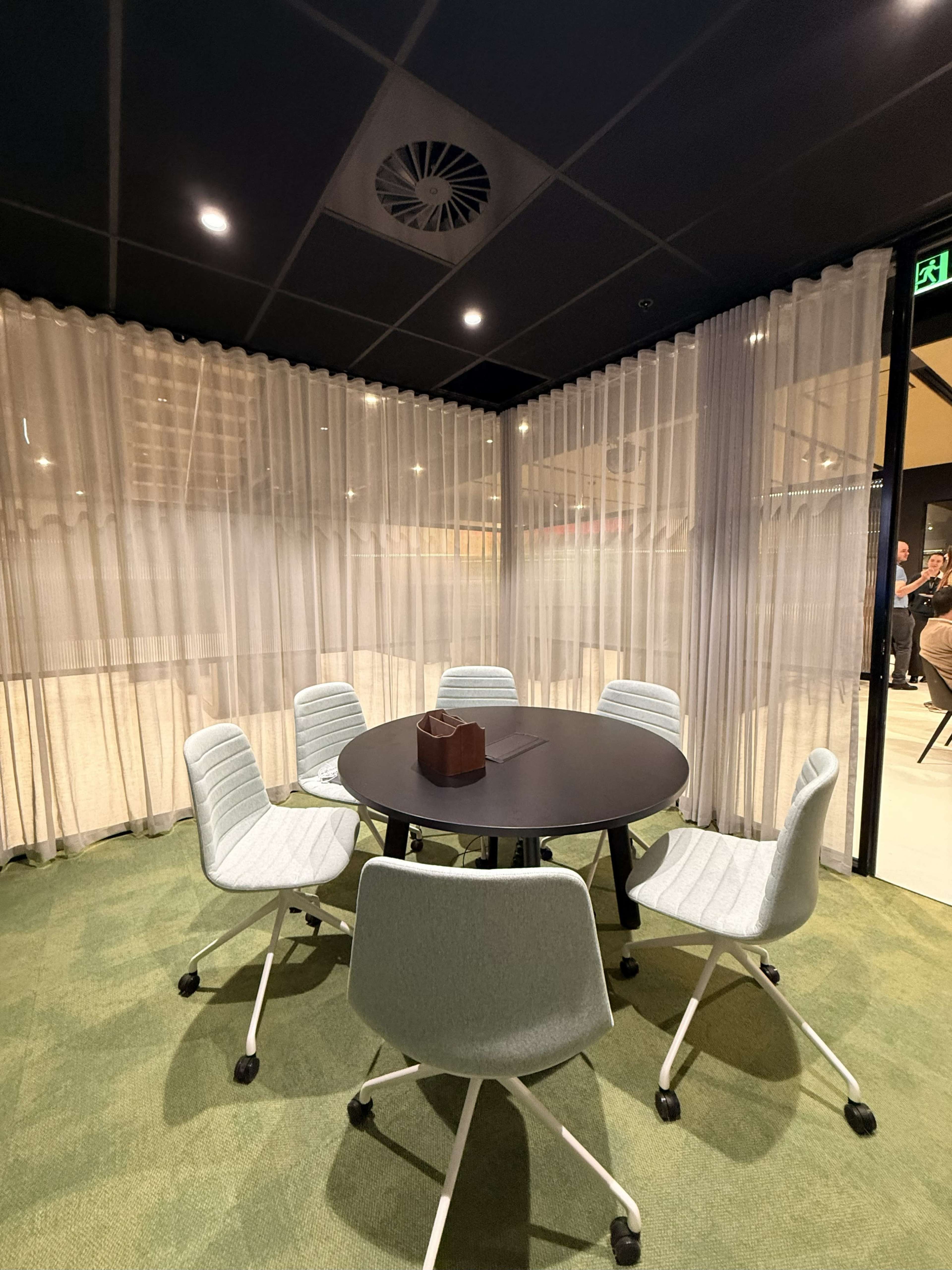 The image shows a small meeting room with a round table surrounded by six light-colored chairs, enclosed by sheer curtains.
