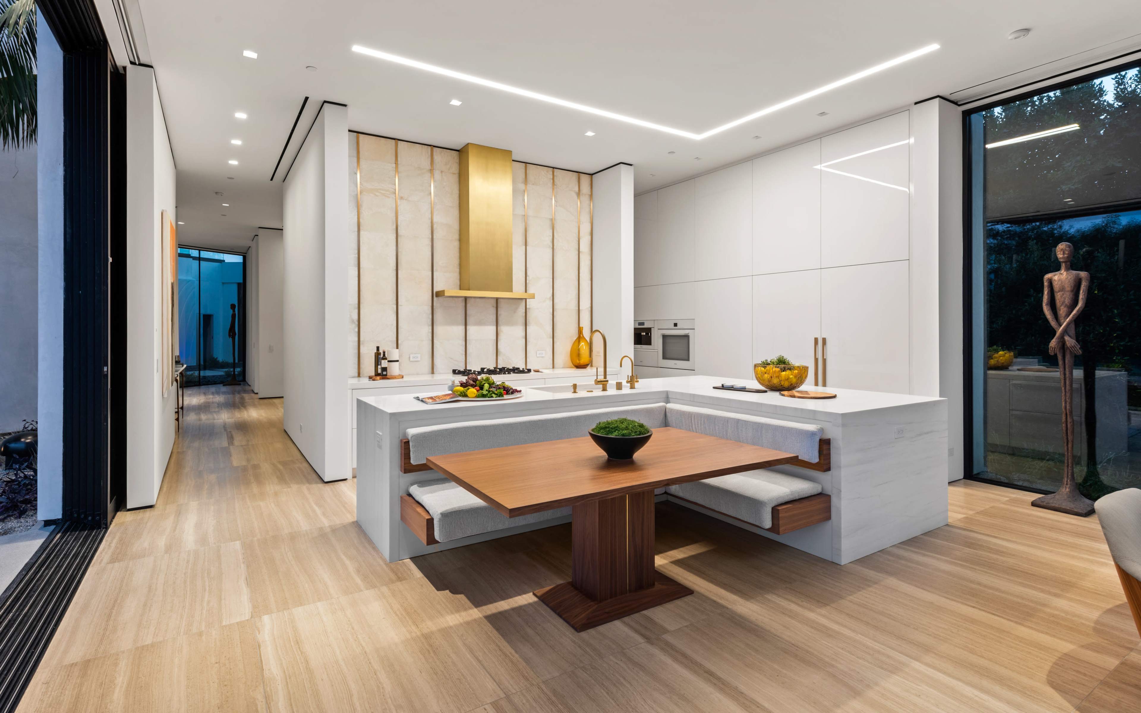 The image shows a modern kitchen with a marble island and a wooden dining table, featuring sleek cabinetry and large windows that allow natural light to fill the space.