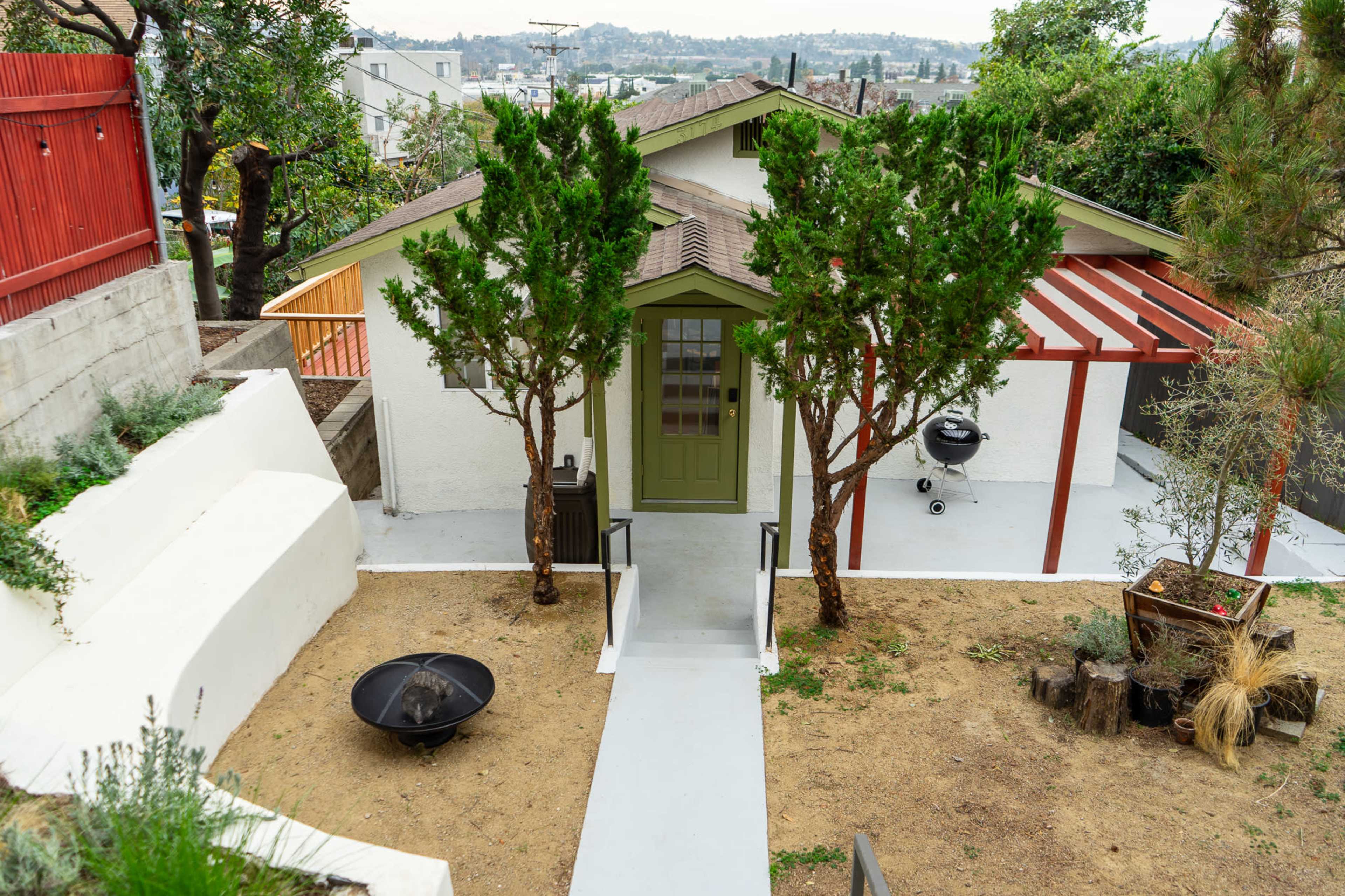 A small white house with a green door is surrounded by two trees and features a patio area with a grill and a fire pit.