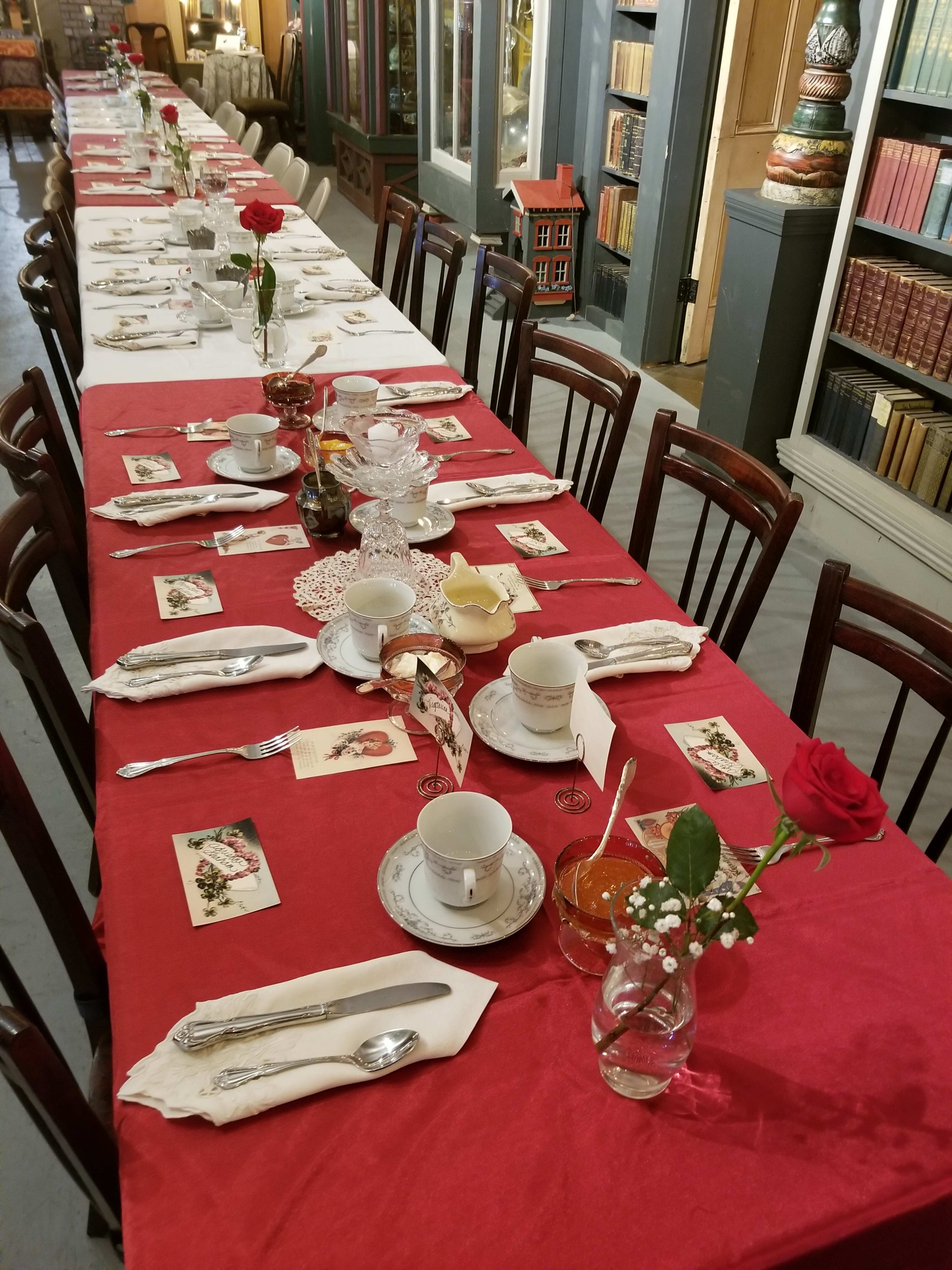 A long dining table is elegantly set with white china, silverware, and decorative flowers, all arranged on a red tablecloth.