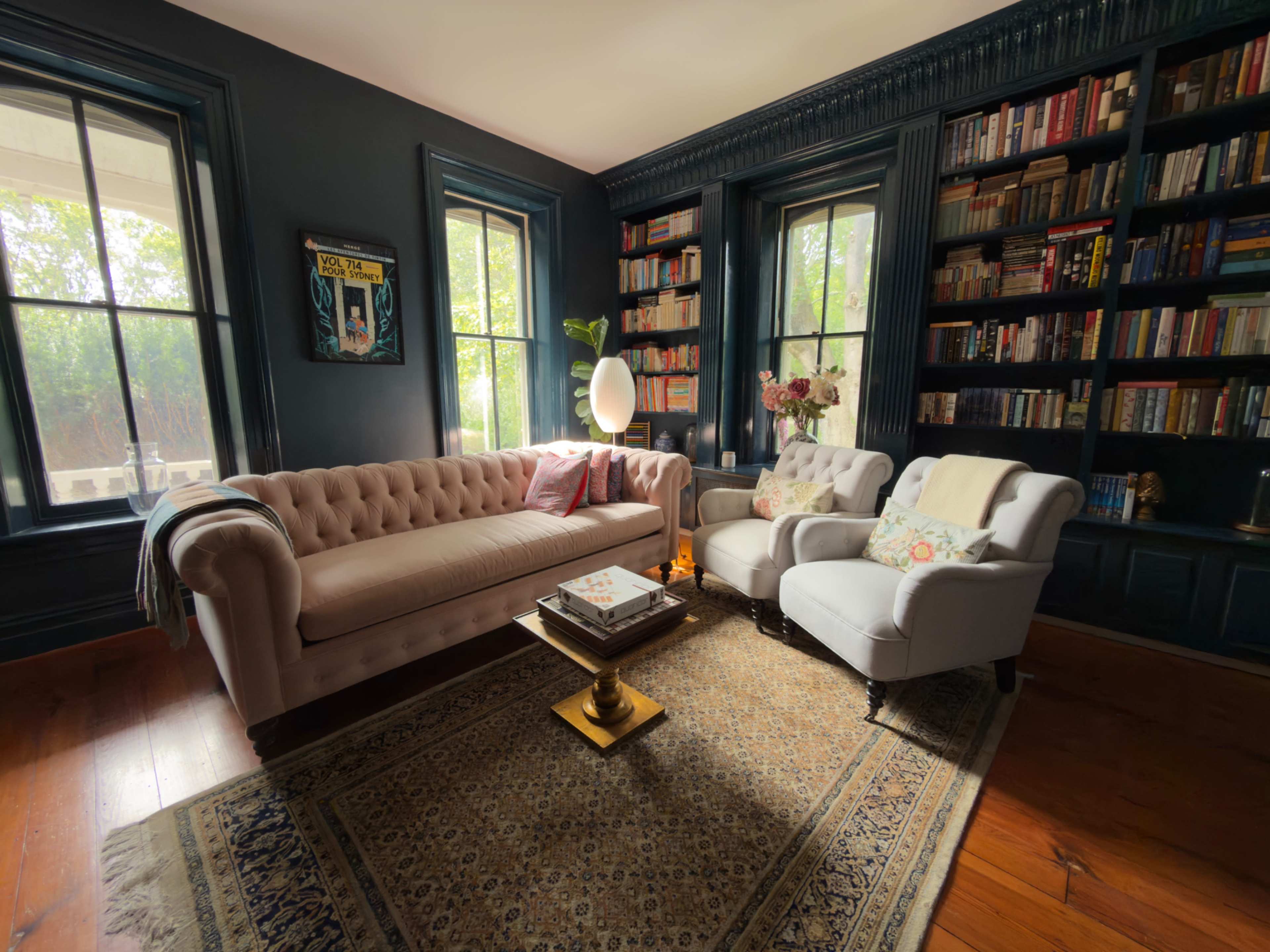 A cozy living room features a tufted pink sofa and a white armchair, surrounded by shelves filled with books and soft natural light streaming through the windows.