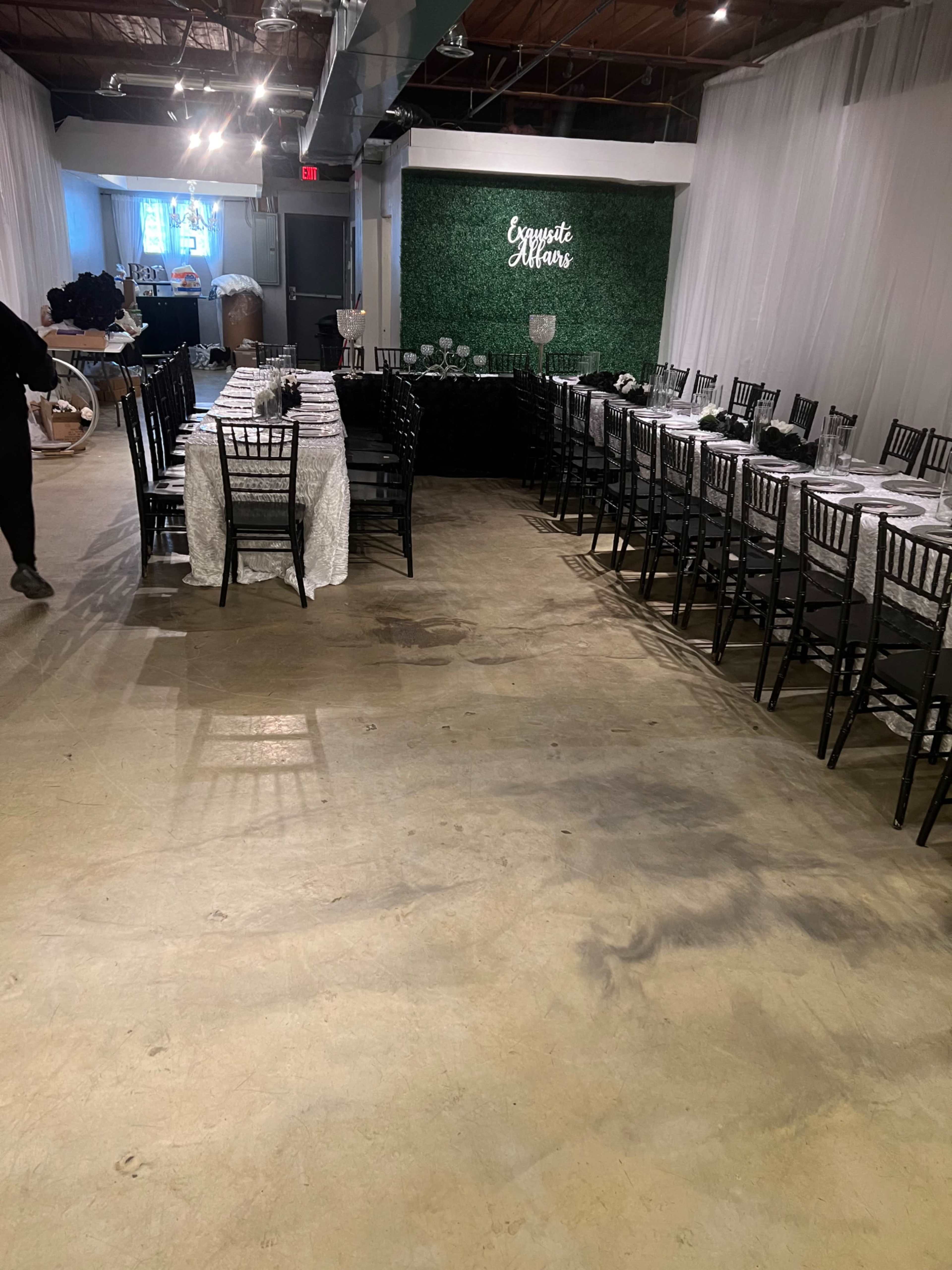 A banquet hall set up with tables covered in white and black linens and black chairs arranged for an event, with a decorative wall in the background.