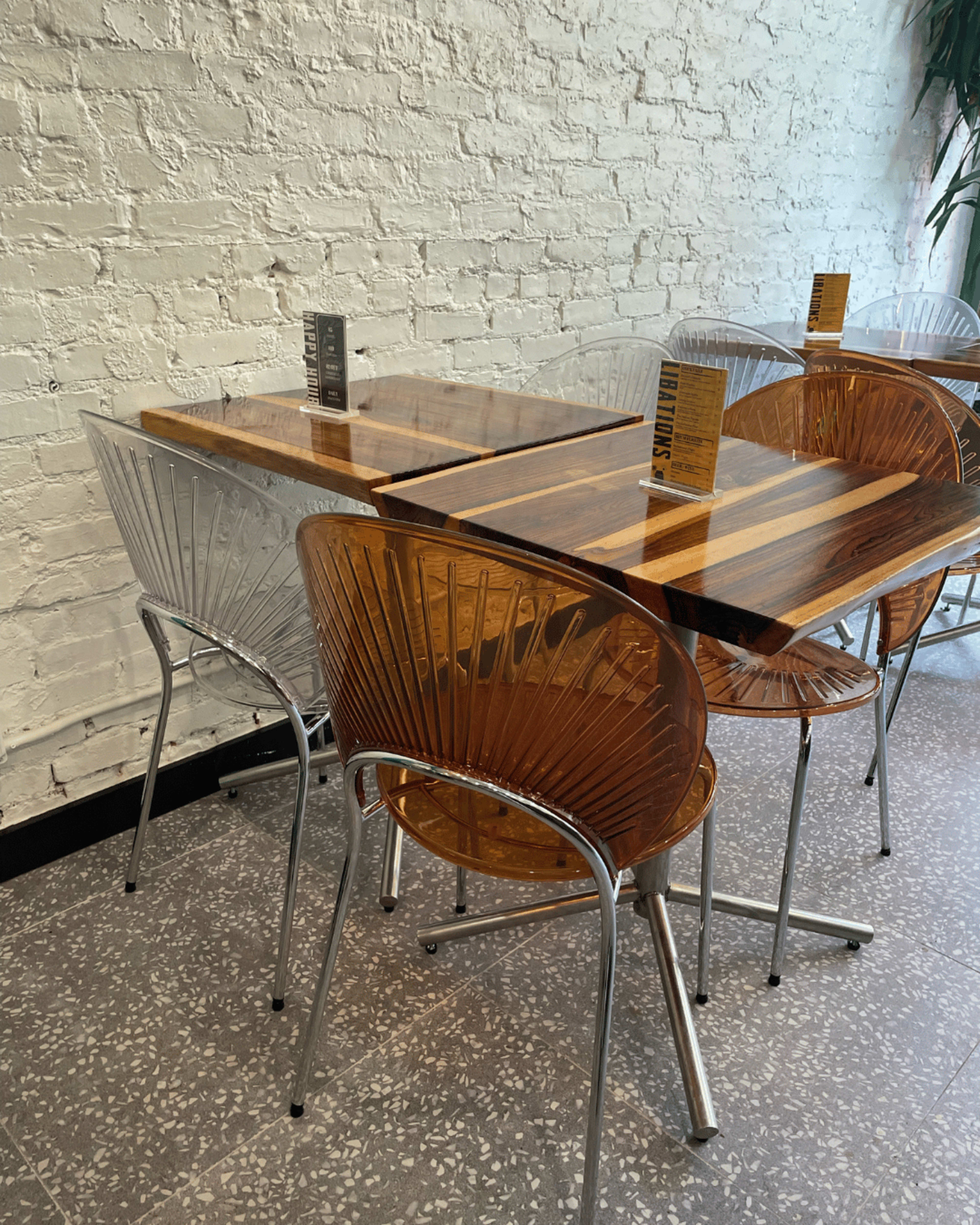 A group of clear and amber acrylic chairs surrounds wooden tables in a dining area with a white brick wall.