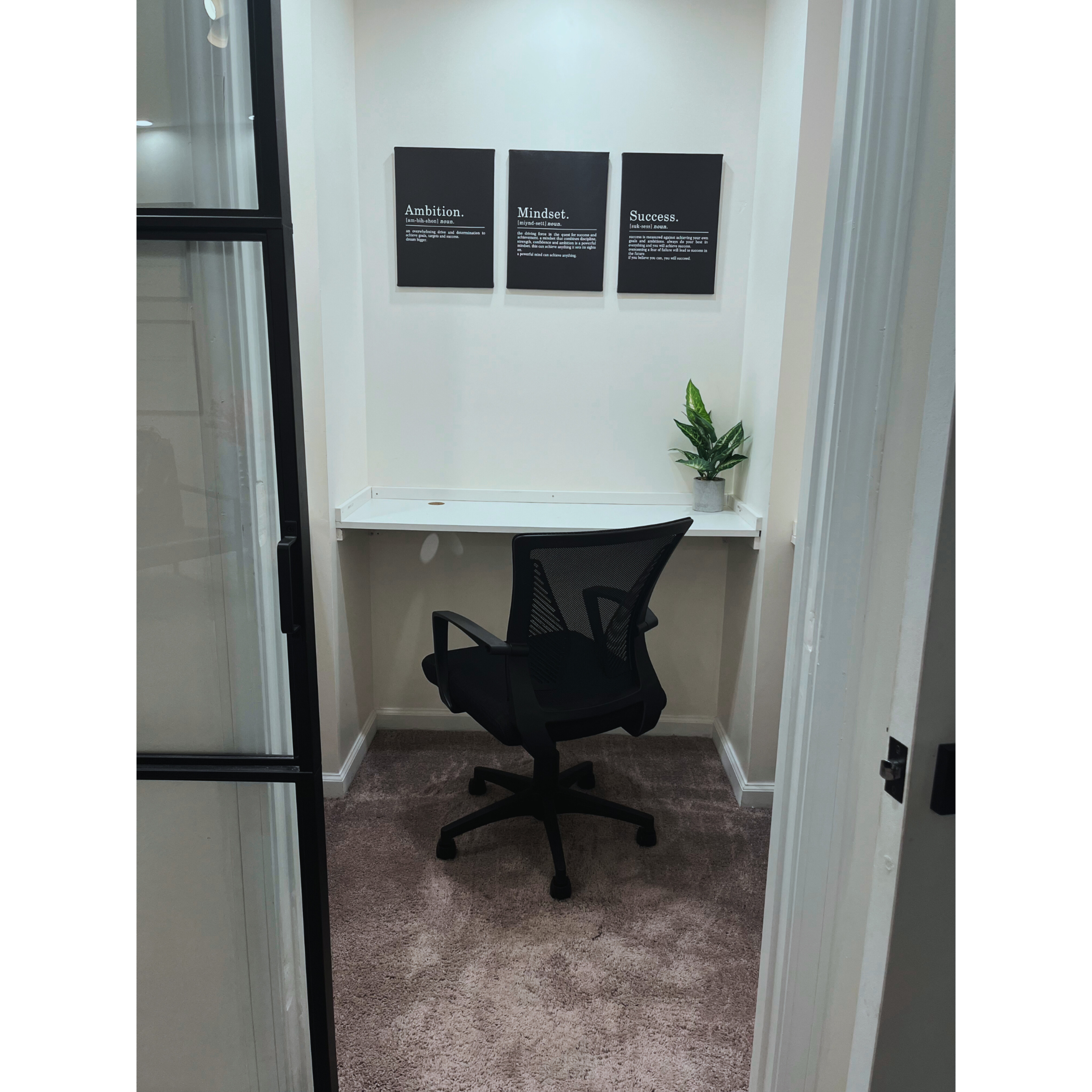 A small office space features a desk with an ergonomic black chair, three framed motivational quotes on the wall, and a potted plant.
