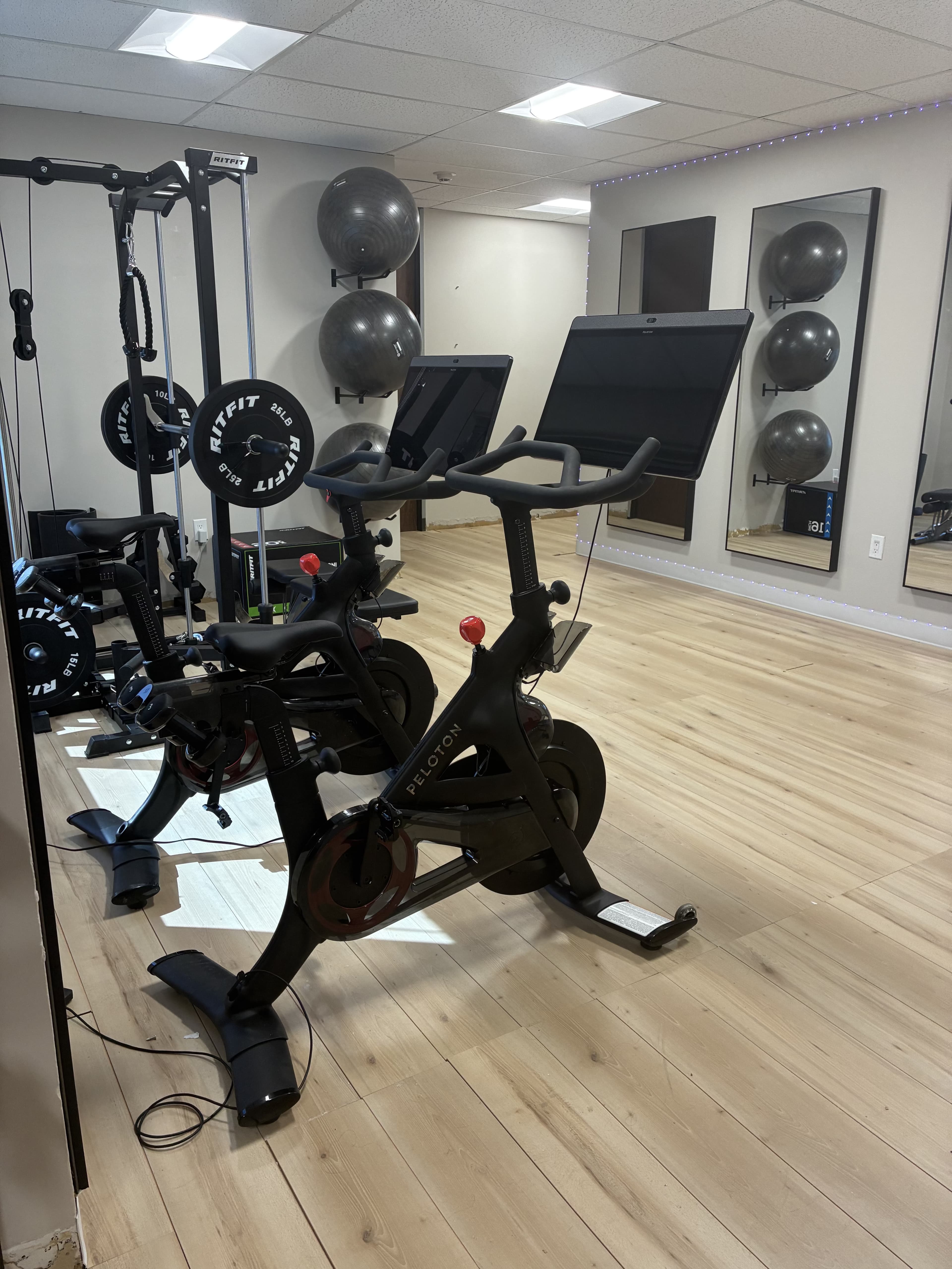 The image shows a fitness studio featuring exercise bikes, weights, and large mirrors in a well-lit room with hardwood flooring.