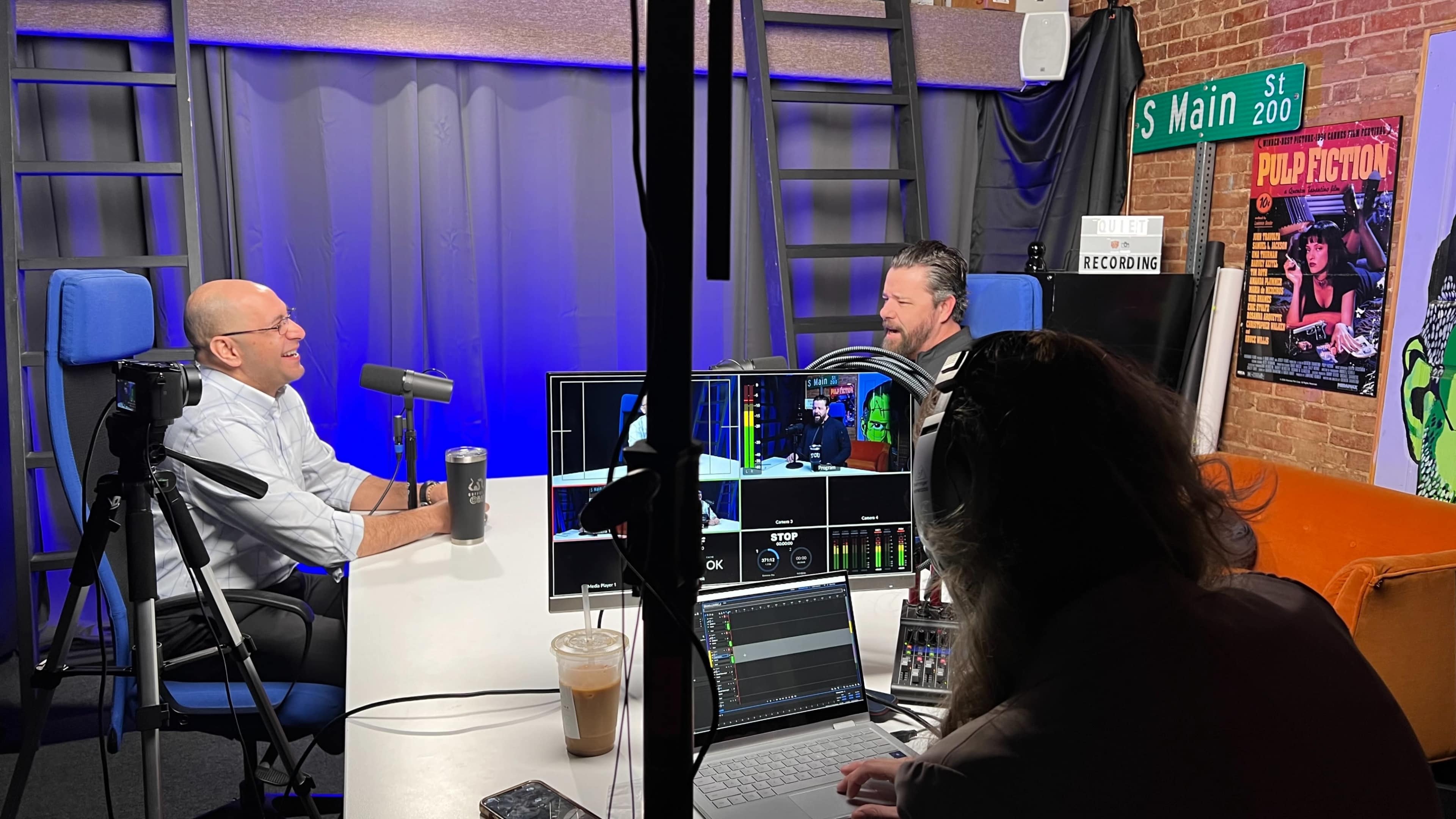 Two men are seated at a table with microphones in a recording studio, while a third person operates equipment and monitors the recording process.