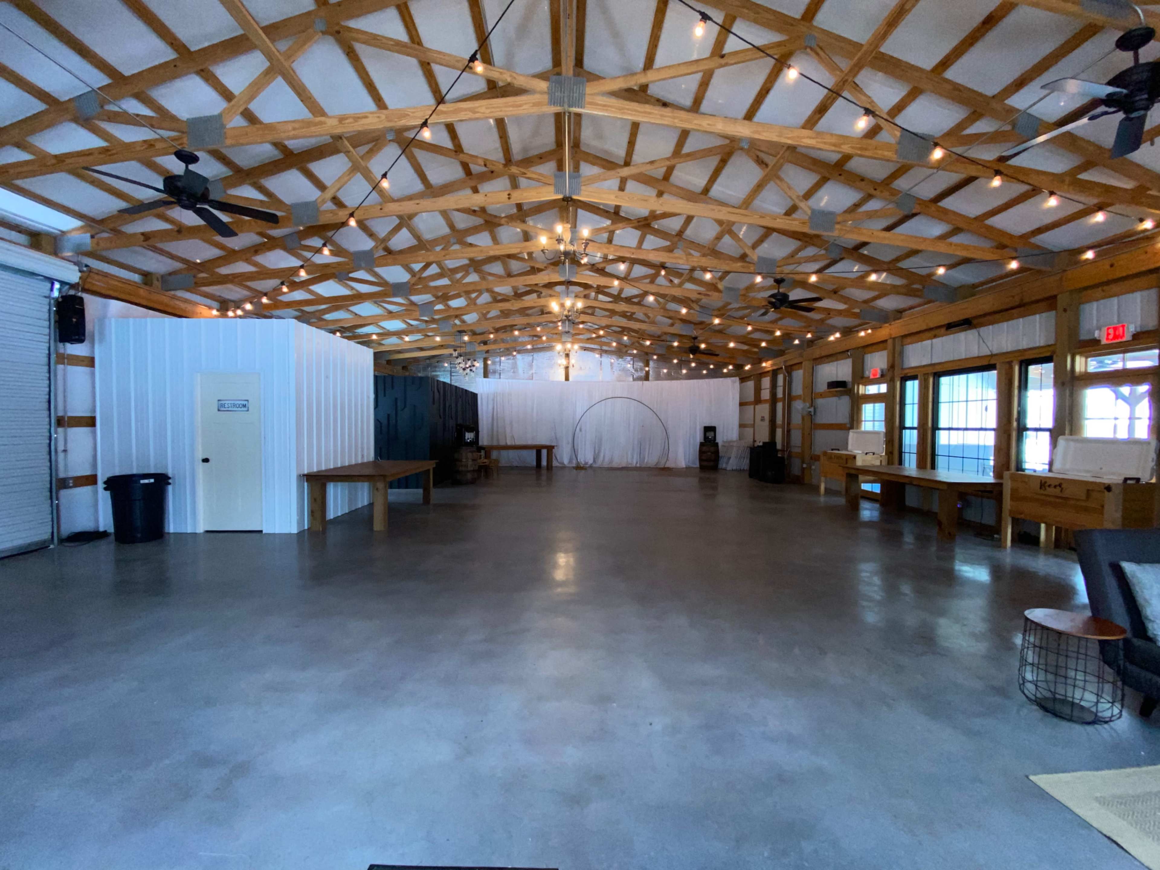The image shows an empty, spacious indoor venue with wooden beams and string lights overhead, featuring concrete flooring and several tables along the walls.