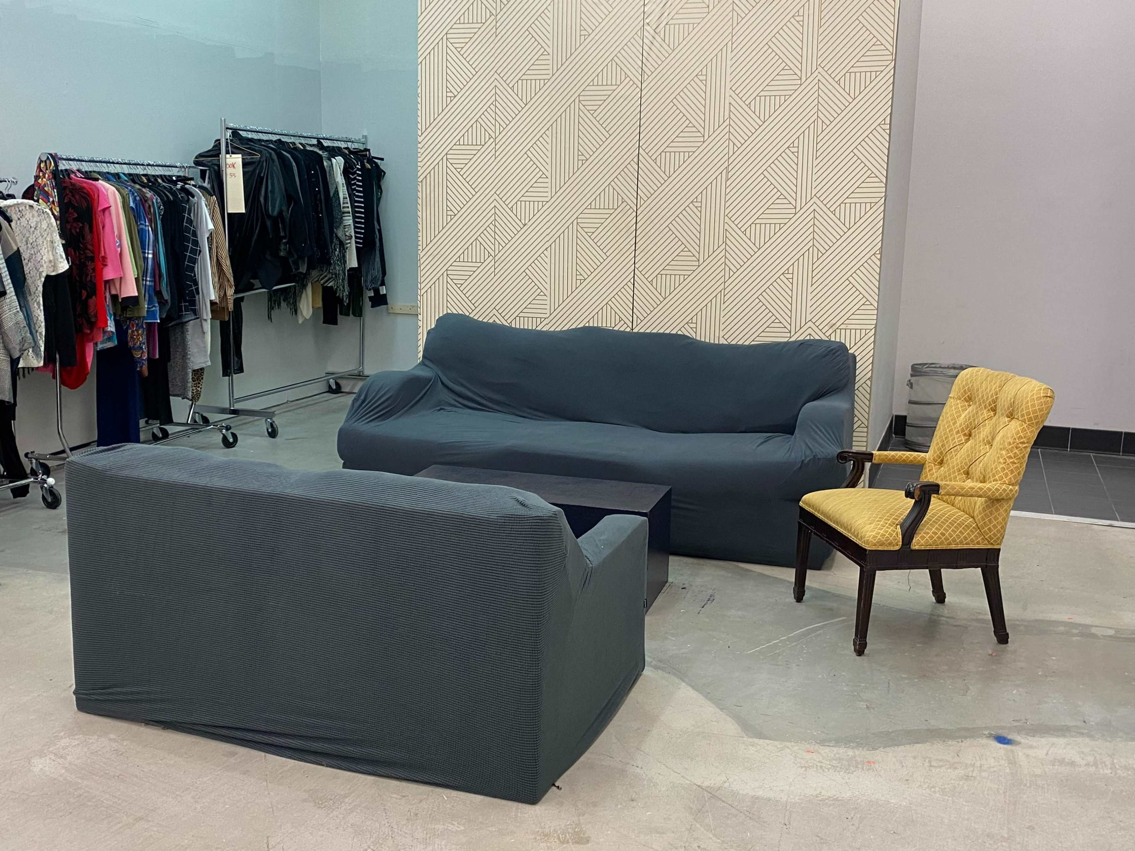 A simple seating arrangement with a gray sofa, a matching loveseat, and a yellow upholstered chair in a room with clothing racks in the background.