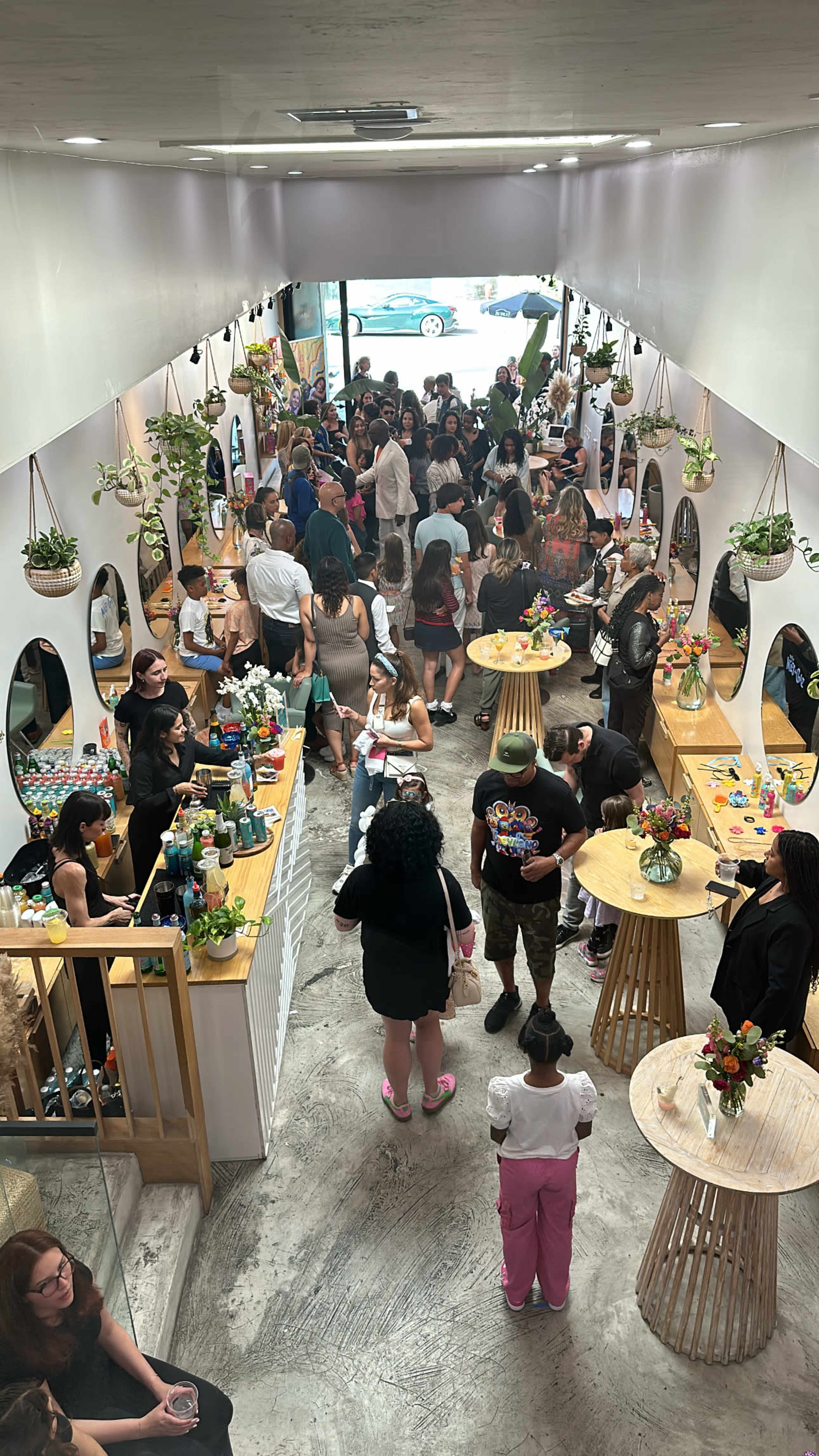 The image shows a crowded indoor space with people socializing around tables adorned with flowers and drinks.