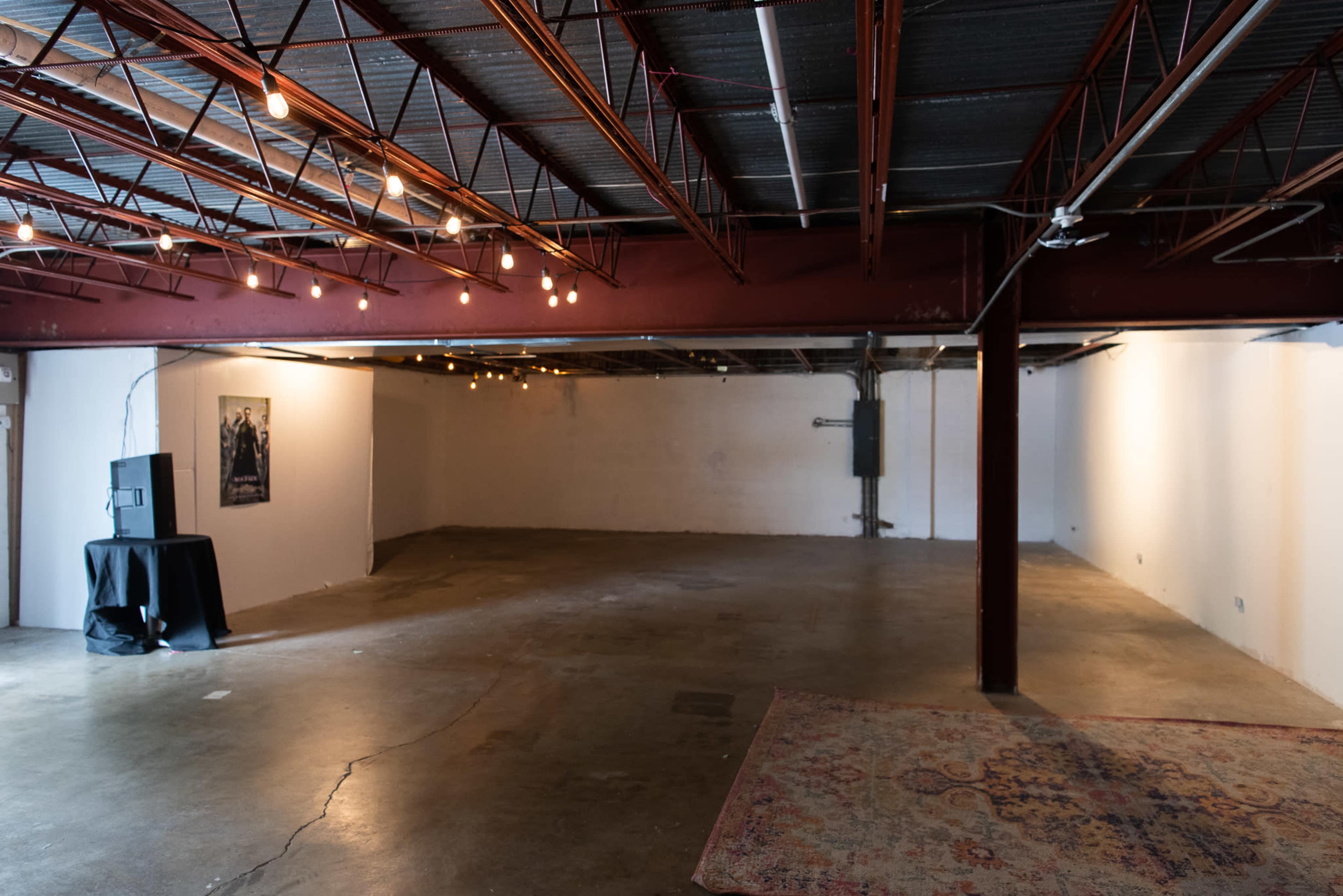 A sparse, industrial-style interior space with exposed beams, concrete flooring, and string lights hanging from the ceiling.