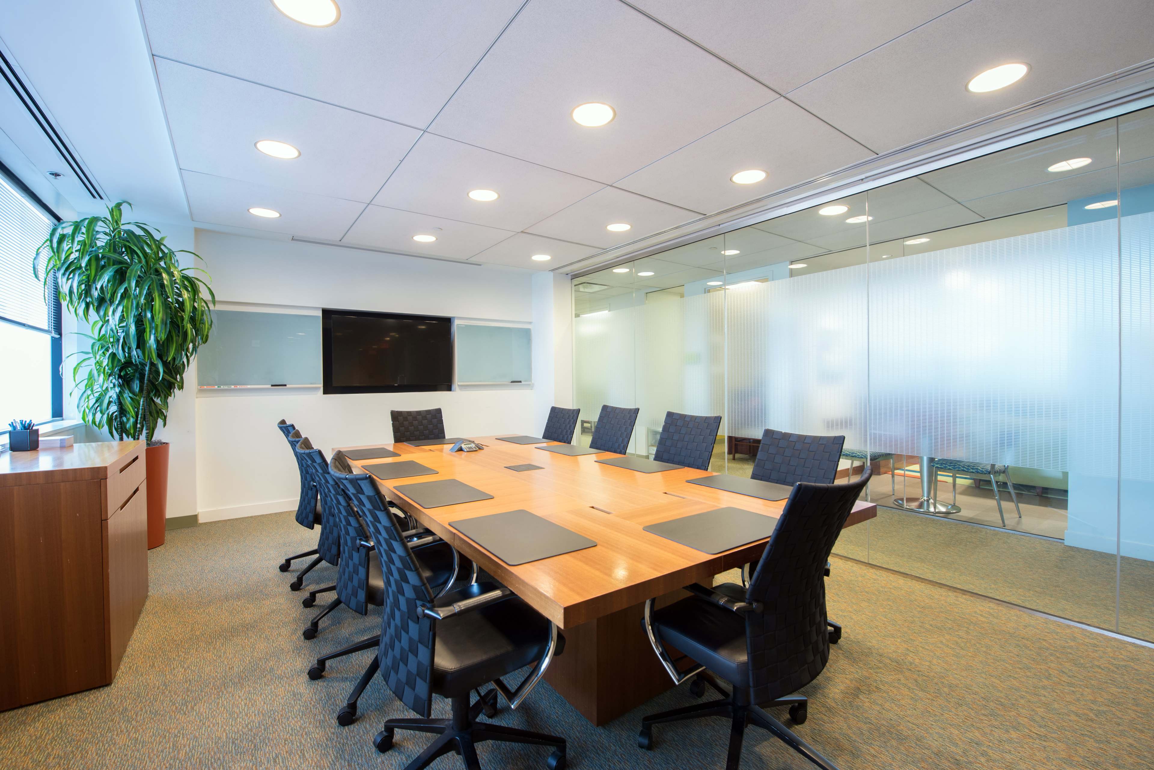 A modern conference room features a large wooden table surrounded by black chairs, with a glass wall and a wall-mounted screen.