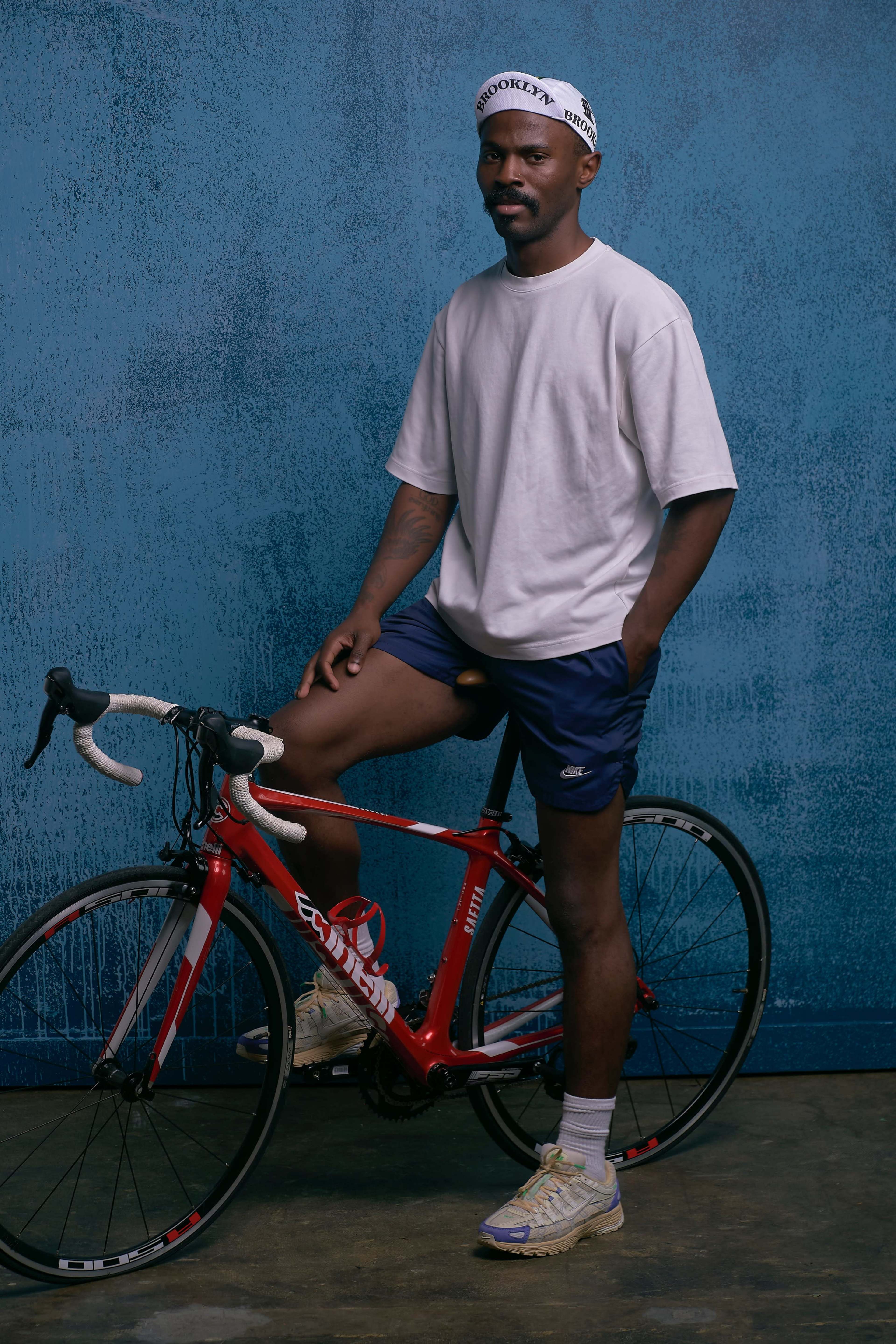 A man in athletic clothing poses next to a red bicycle against a blue textured wall.