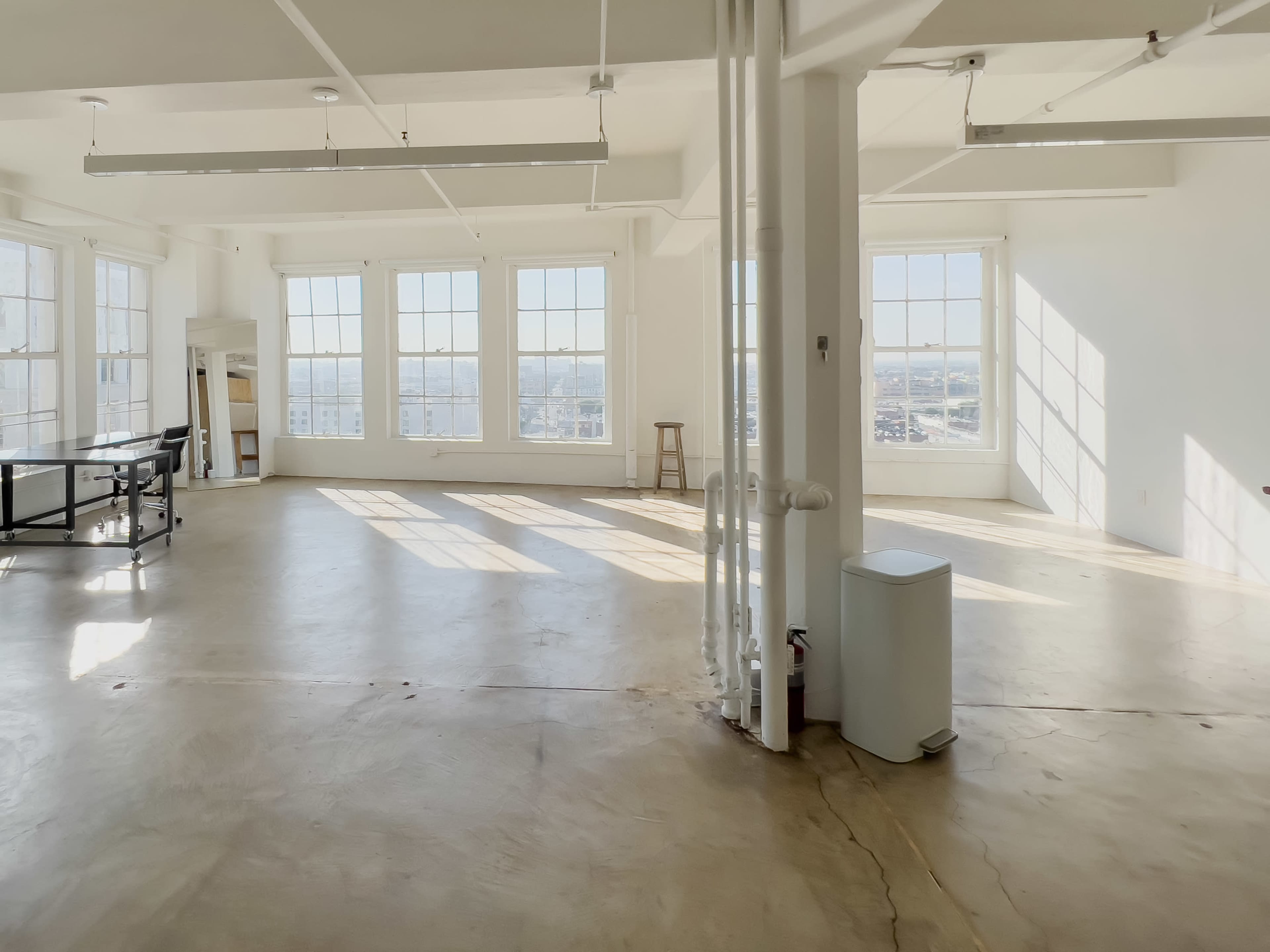 The image shows a spacious, sunlit room with large windows, concrete flooring, and minimal furnishings.