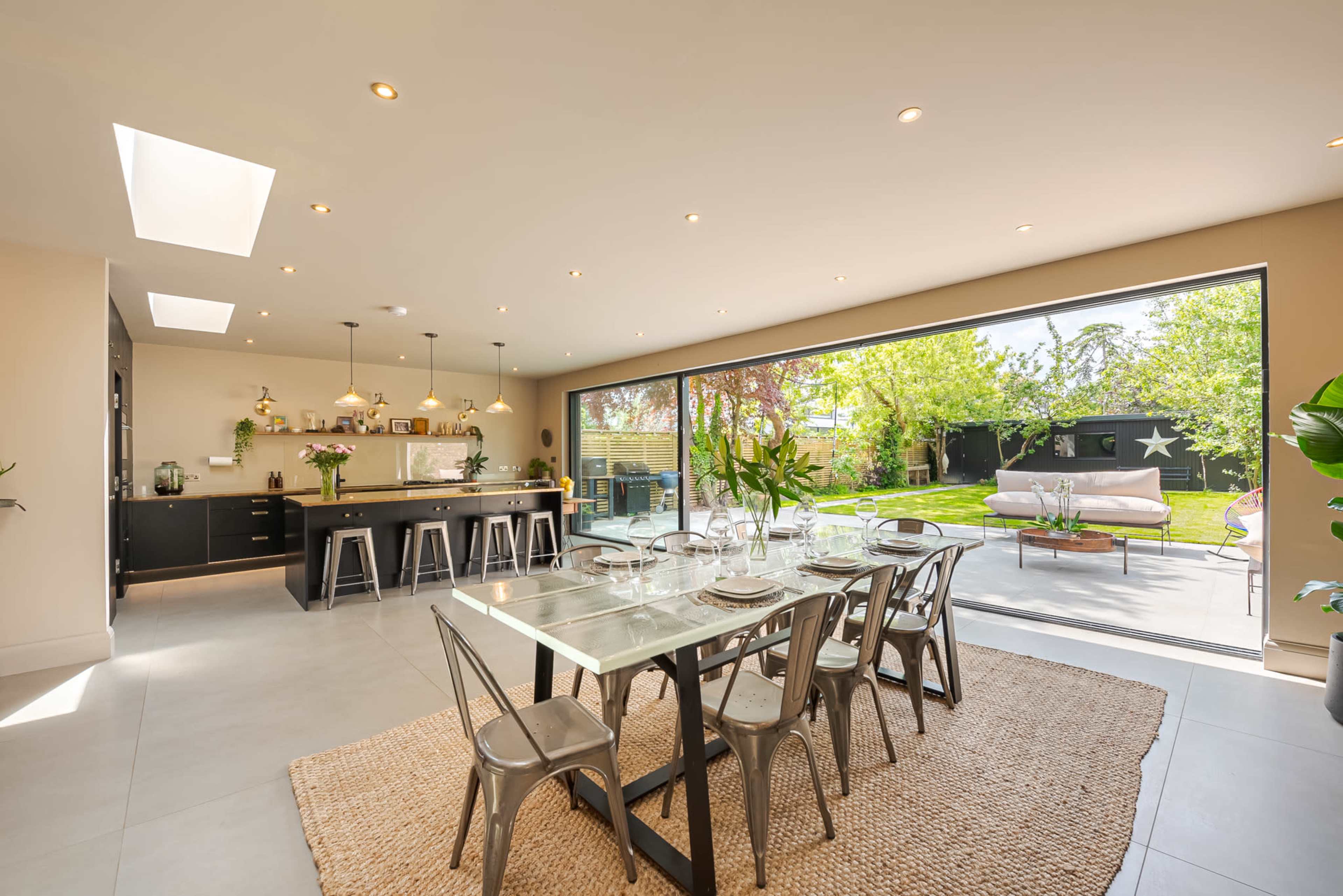 A modern dining area with a glass table set for a meal, adjacent to an open kitchen and overlooking a spacious garden through glass sliding doors.