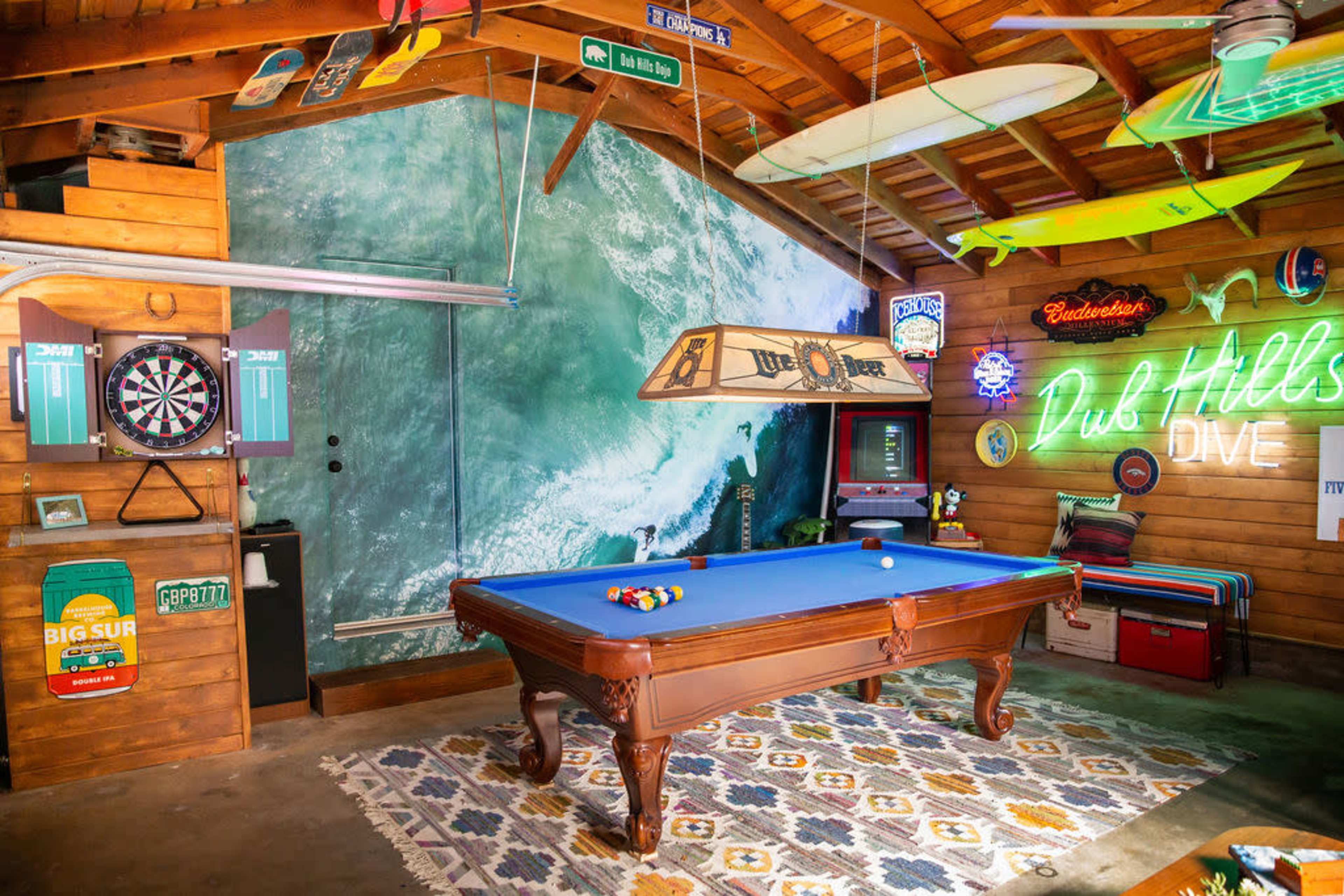 A game room featuring a blue billiard table, a dartboard, and neon signs, with a surfboard and ocean mural on the wall.
