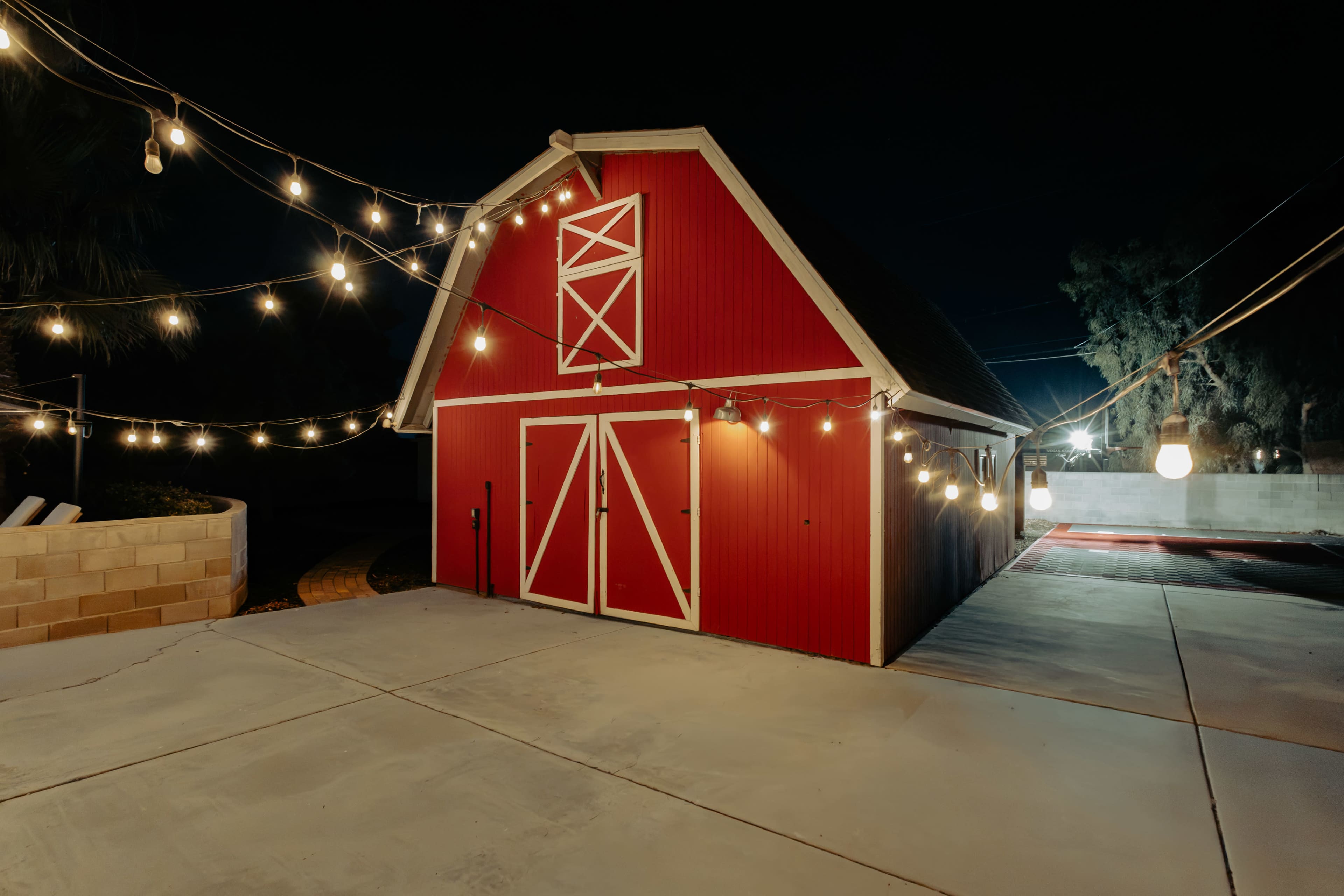 A red barn illuminated by string lights stands in a paved area at night.
