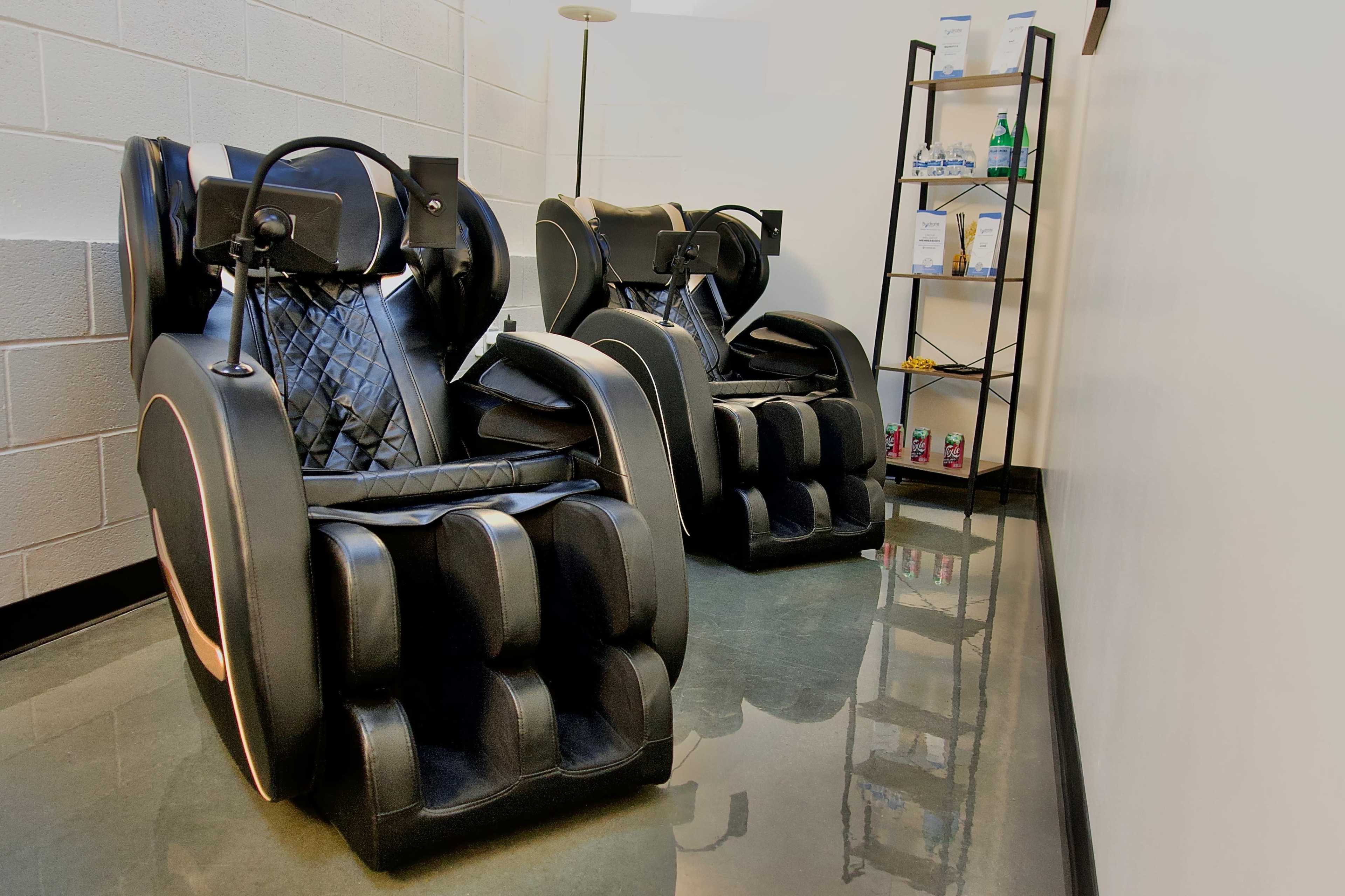 The image shows two black massage chairs positioned against a wall, with a small shelf nearby displaying various bottles and products.