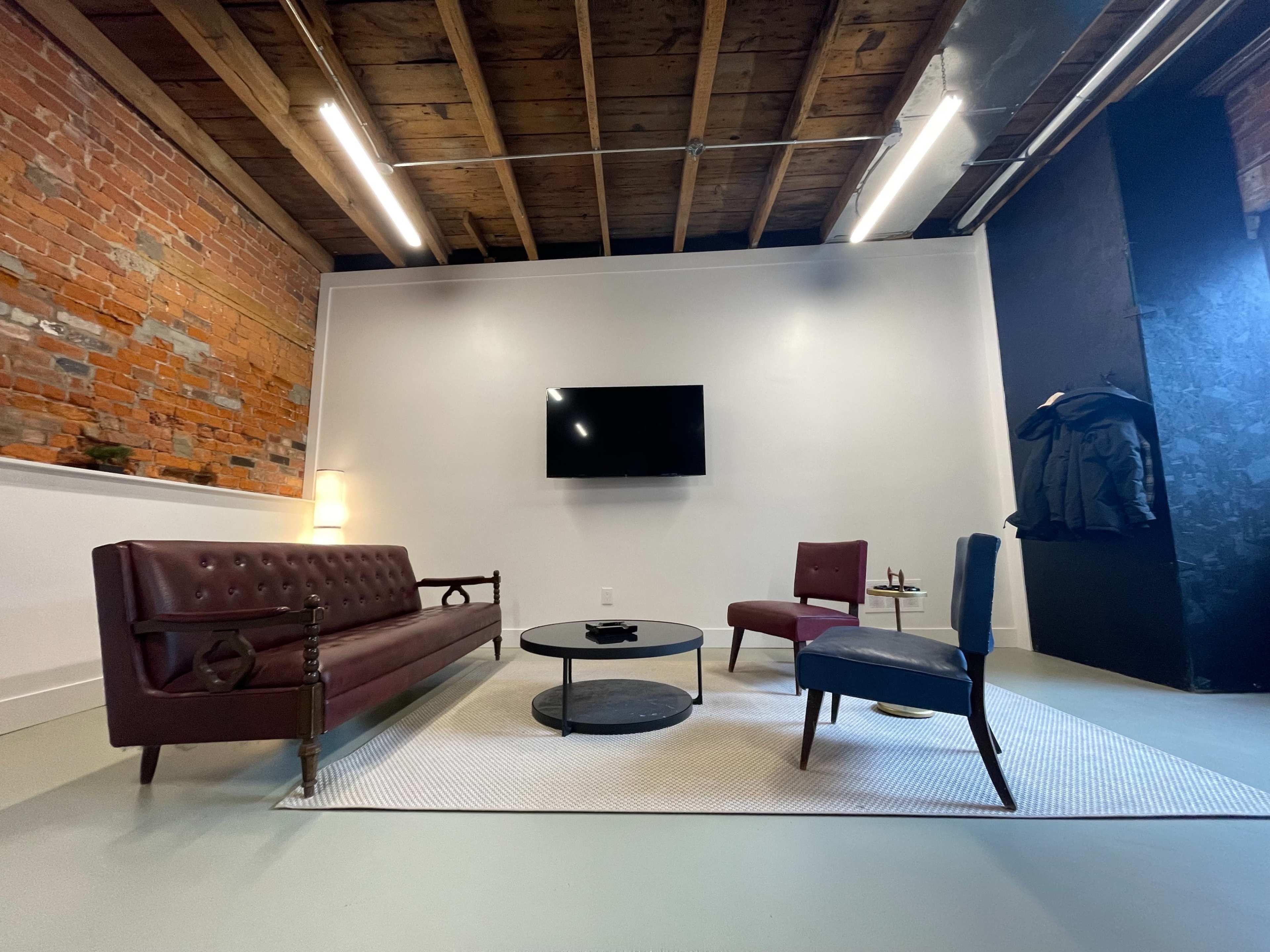 A living room with exposed brick walls, a maroon sofa, a blue chair, a round coffee table, and a television mounted on the wall.