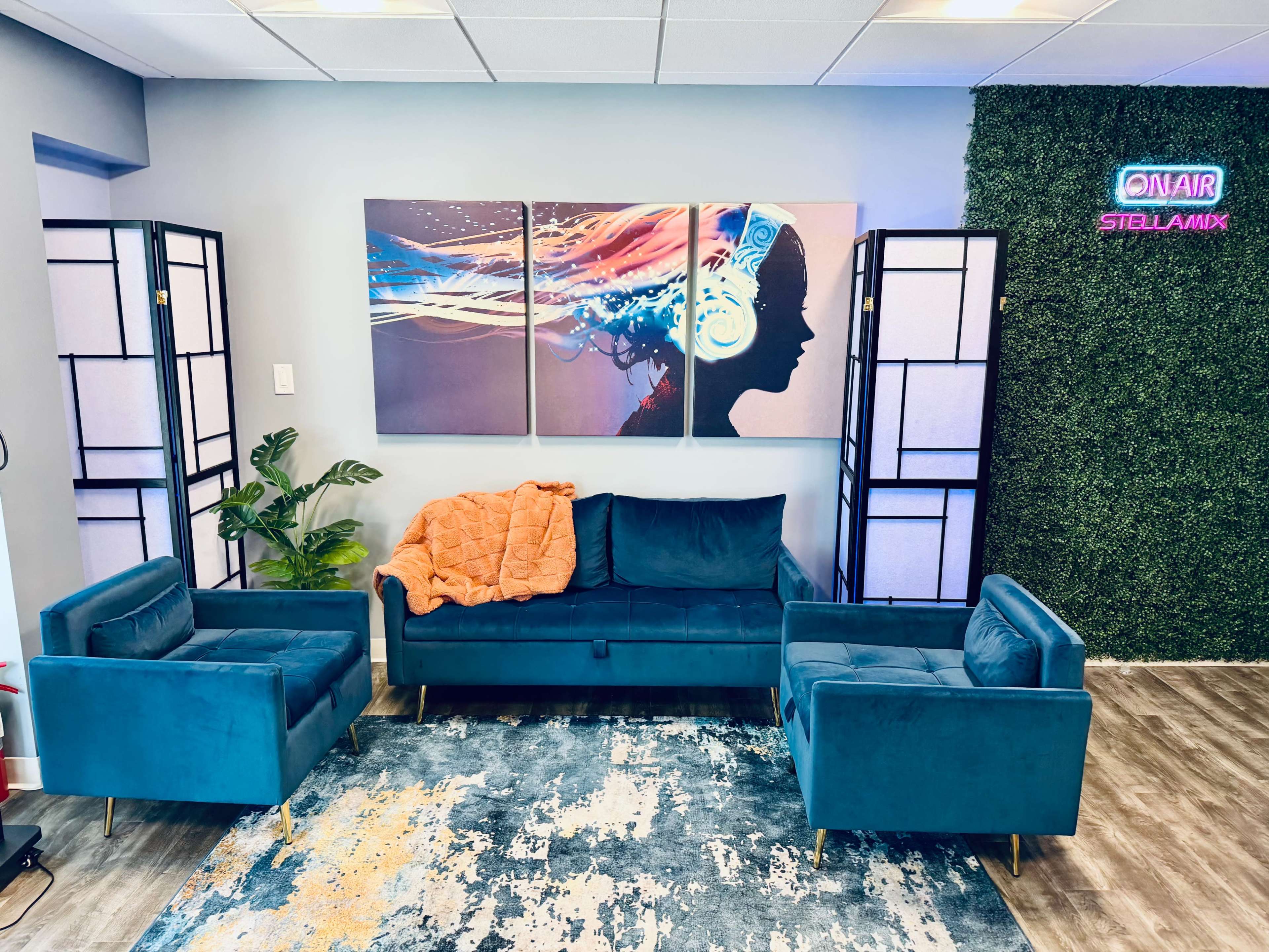 The image features a modern lounge area with a teal sofa and armchairs, a colorful abstract wall art piece, and decorative lighting, all set against a green plant wall.