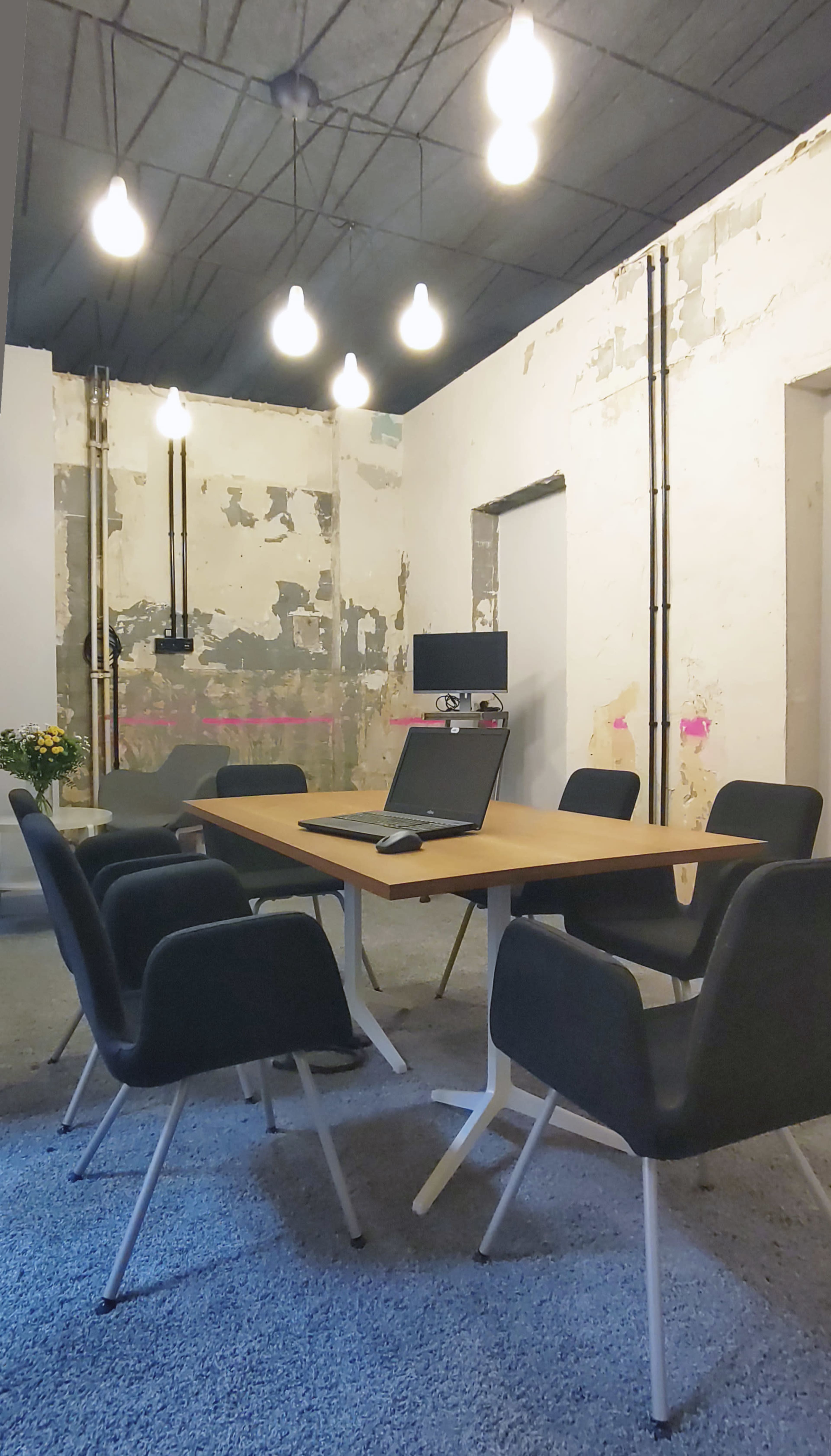 A modern conference room features a wooden table surrounded by black chairs, with exposed walls and overhead lights.