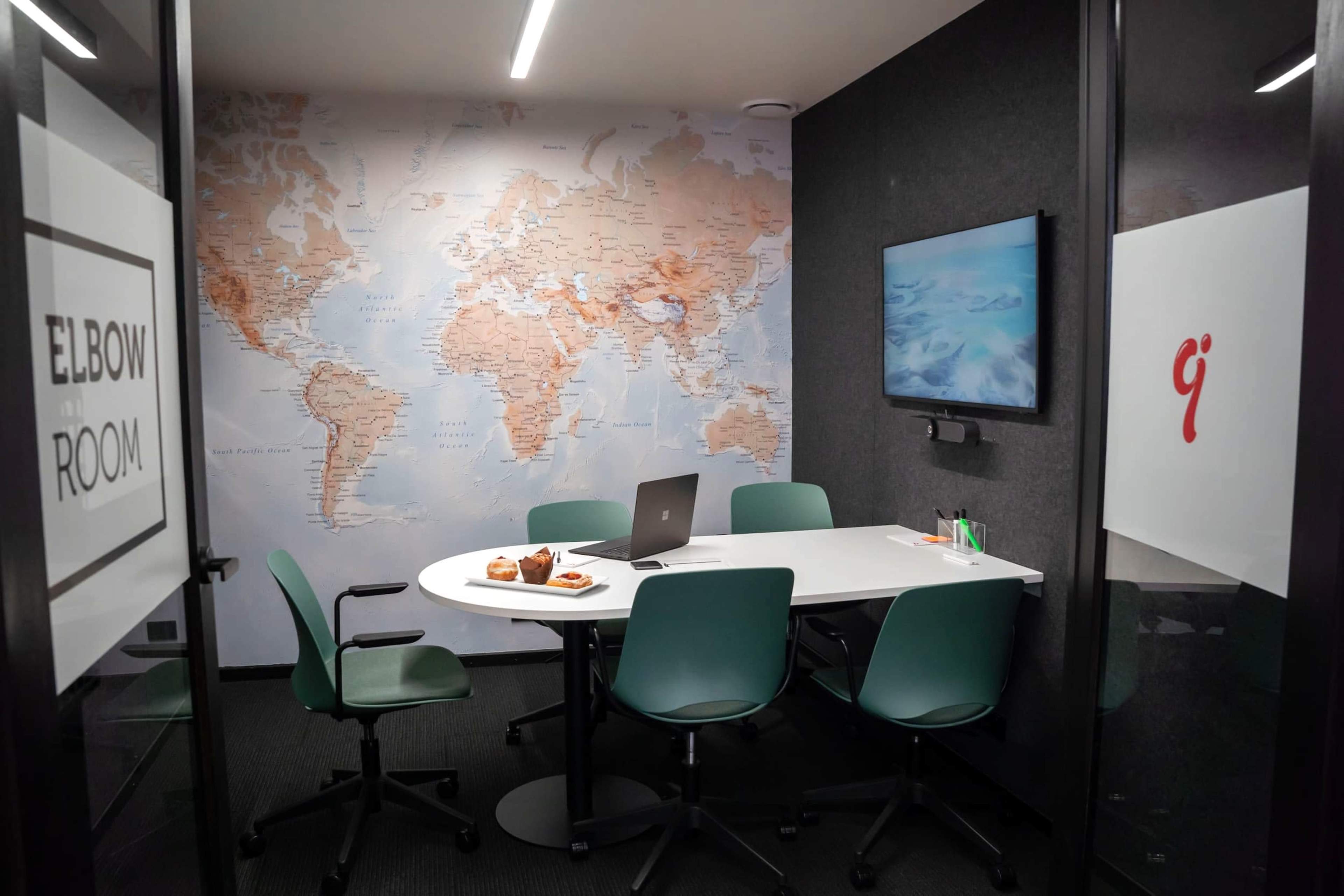 The image shows a small conference room with a white table, green chairs, a laptop, and snacks, featuring a large world map on the wall and a screen displaying content.
