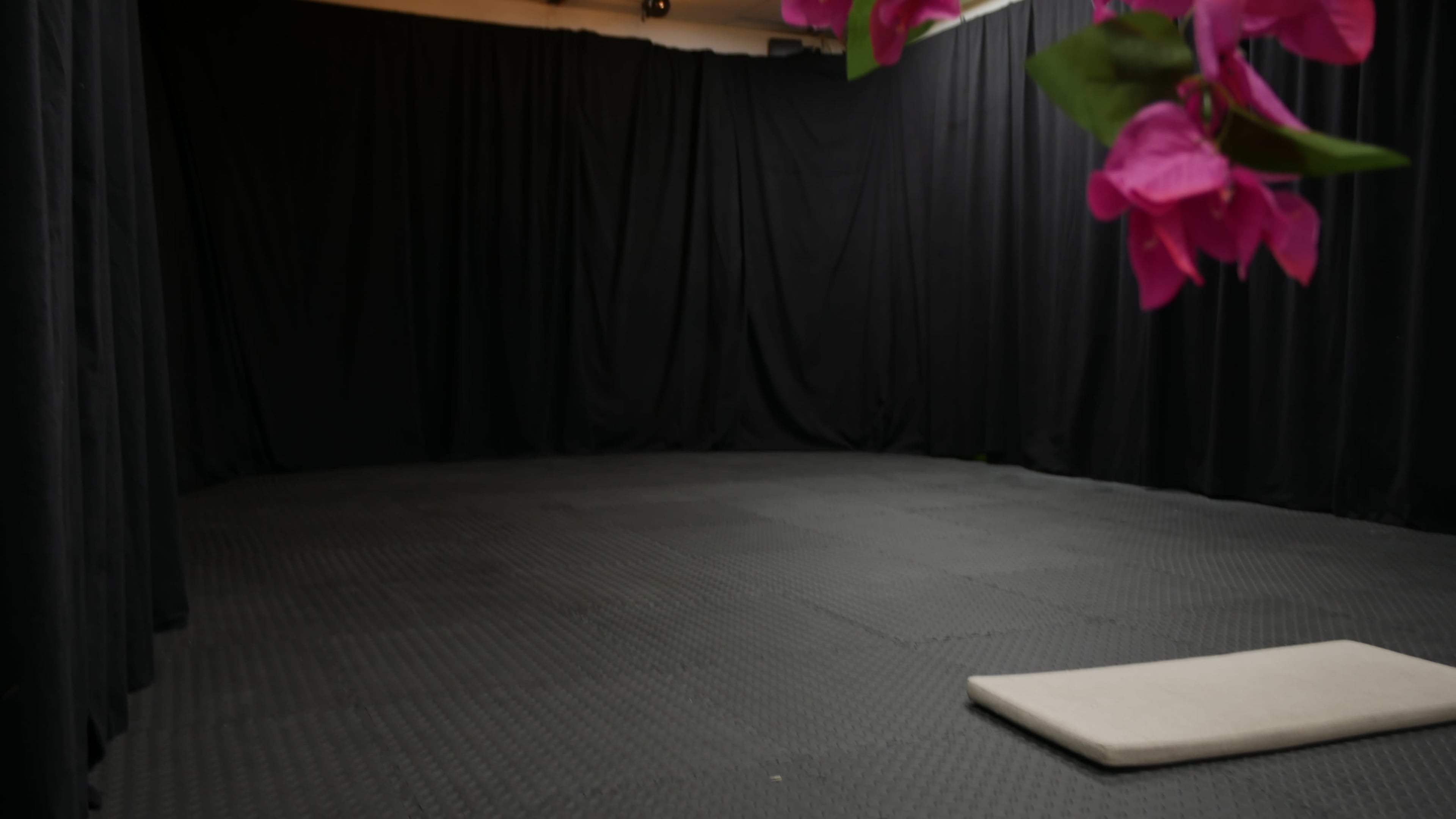 The image shows a dimly lit rehearsal space with black curtains and a small mat positioned on the floor.