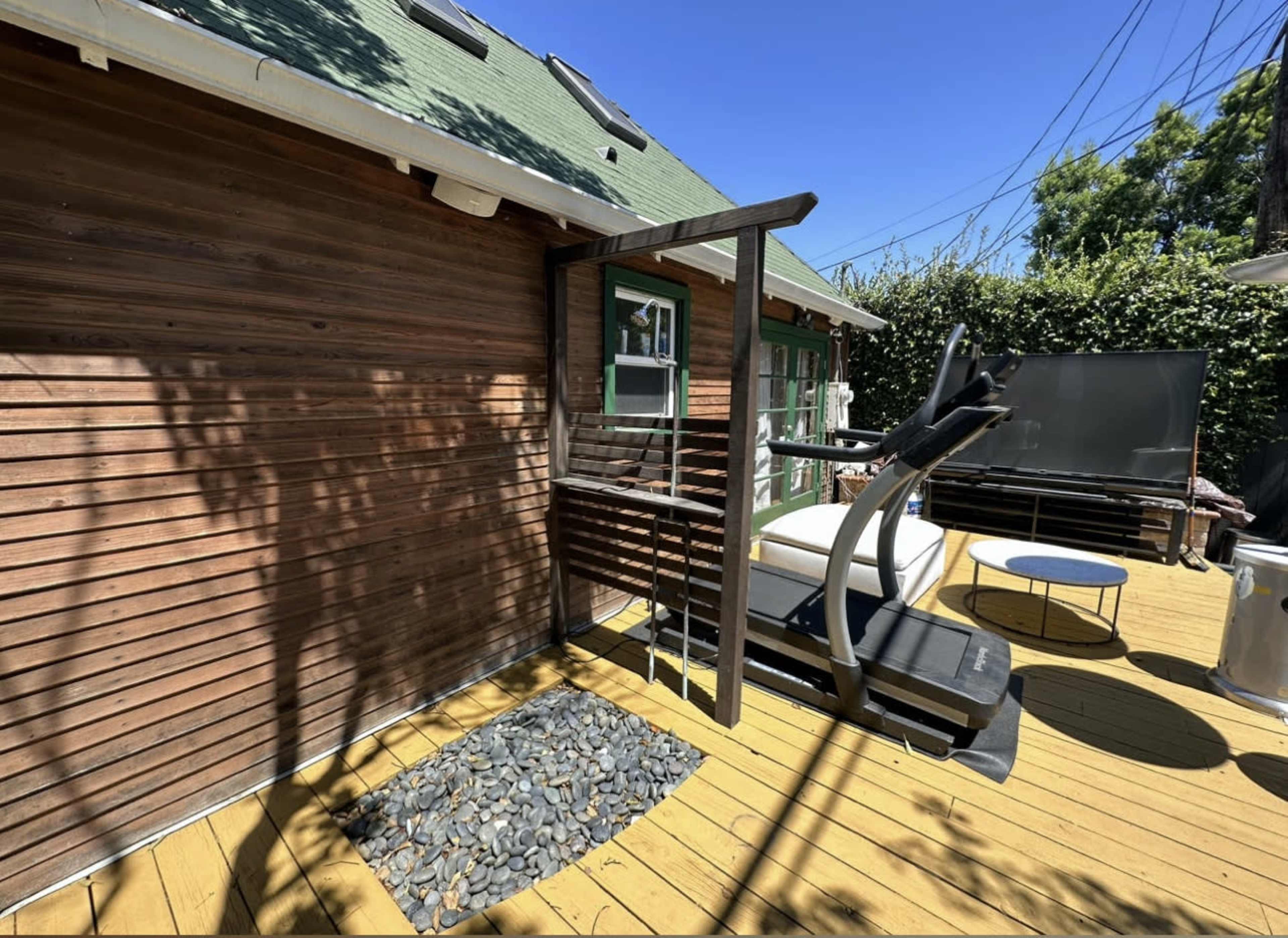 A wooden deck with a fitness machine positioned next to a low table, surrounded by greenery and a clear blue sky.
