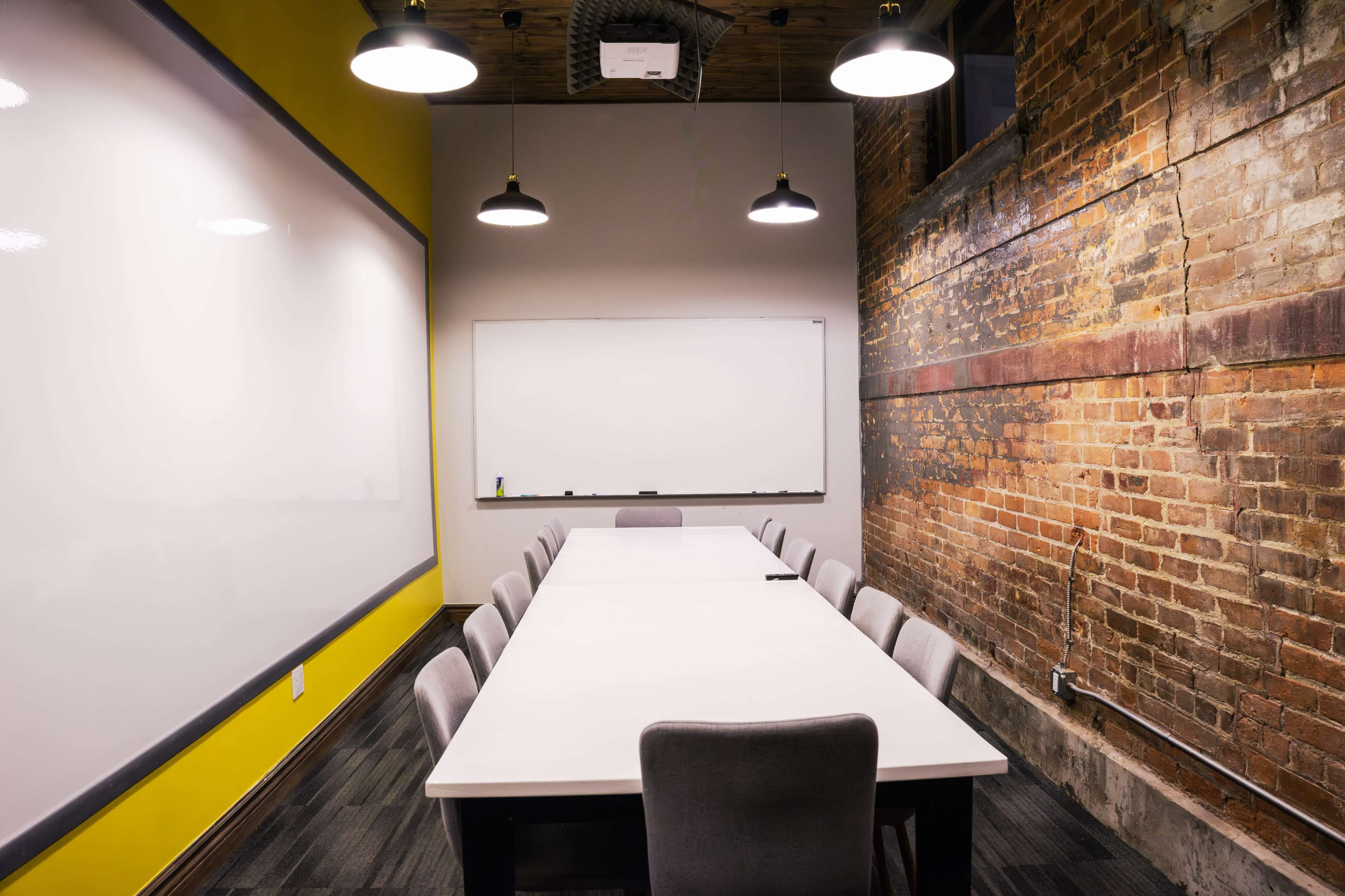 A long table with gray chairs is situated in a conference room featuring a whiteboard and exposed brick walls.