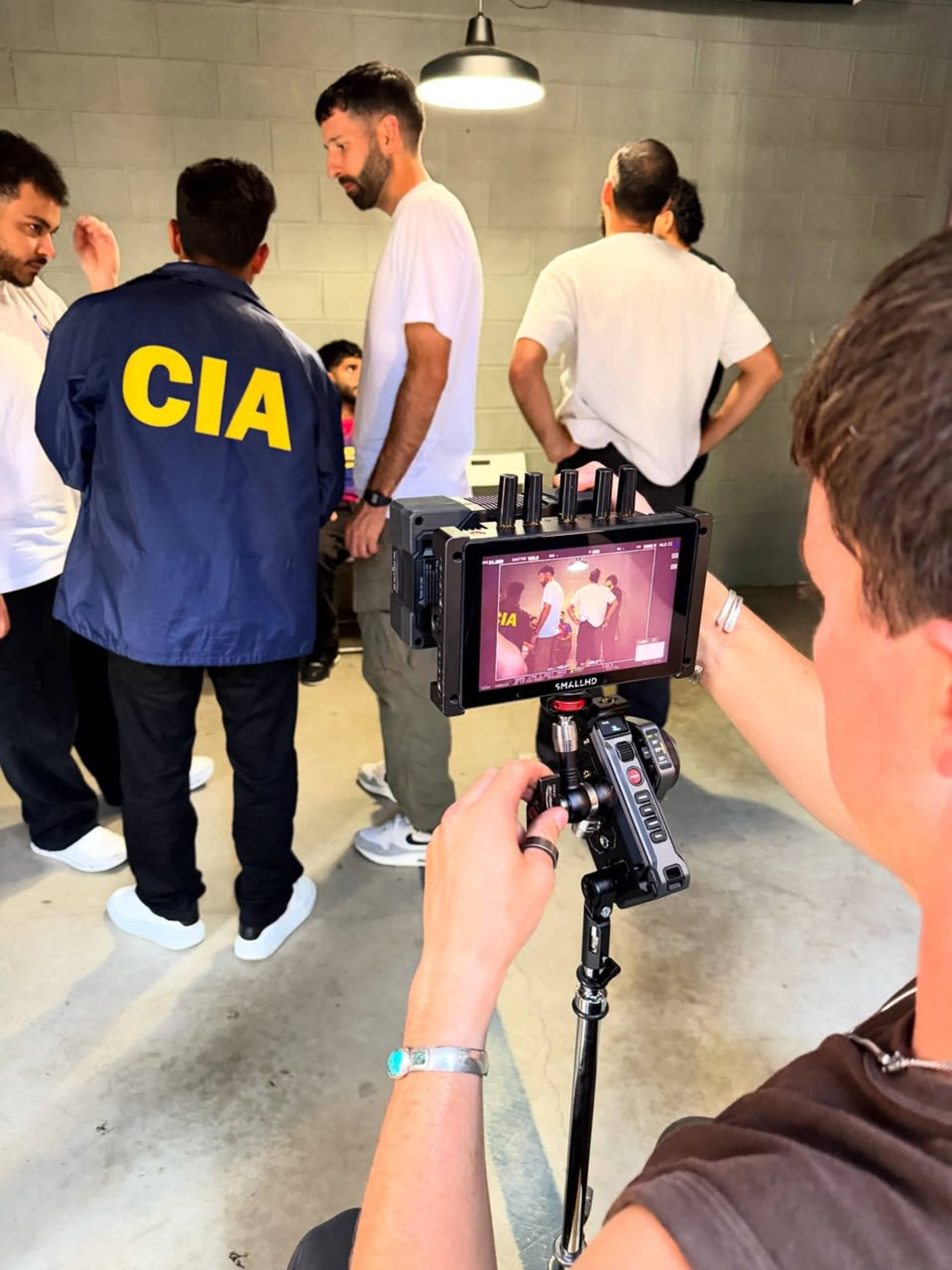 A person operates a camera on a tripod while filming a group of individuals in a gray-walled room.
