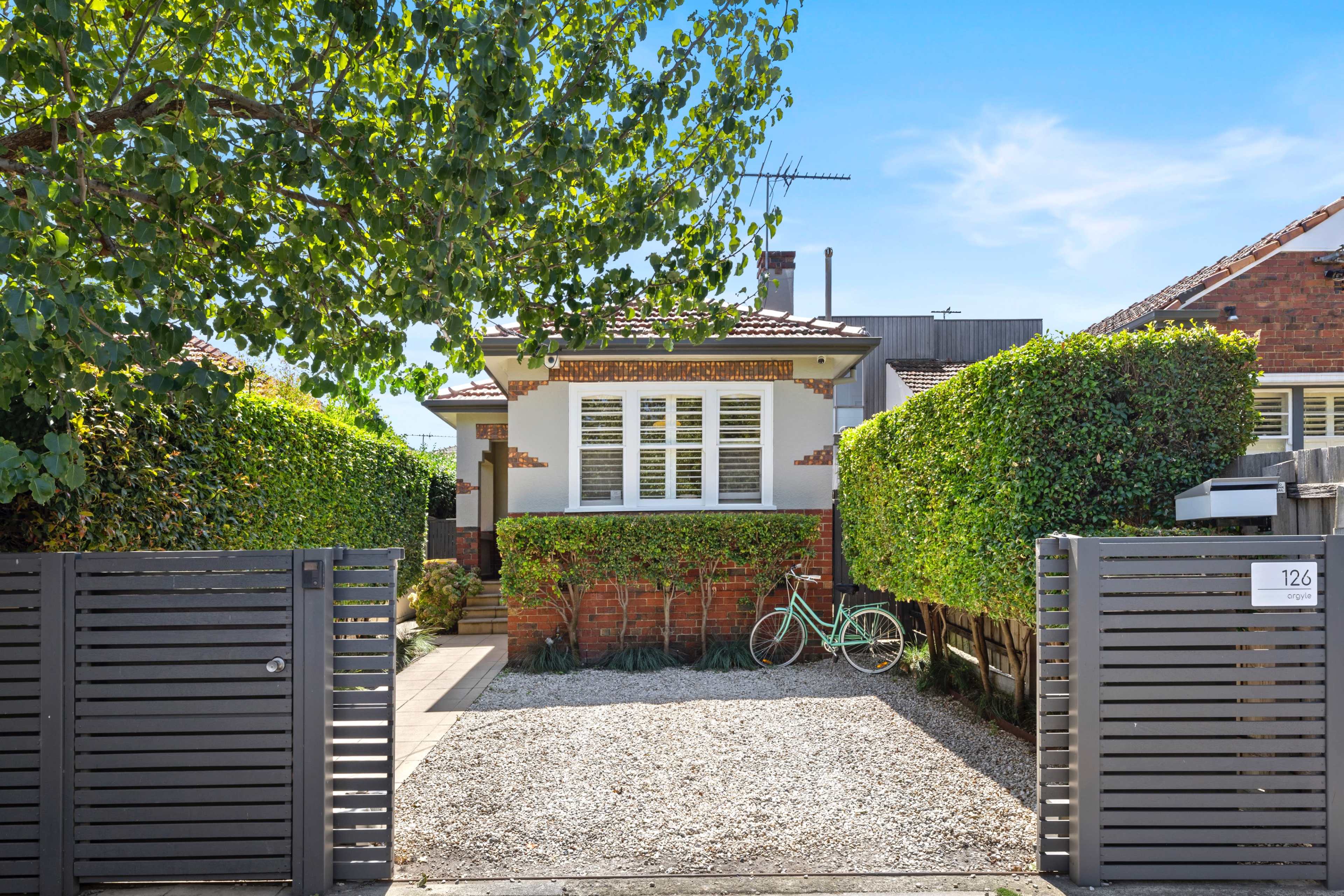 A single-story house with a neatly landscaped front yard and a bicycle parked beside it is enclosed by low hedges and a modern gate.