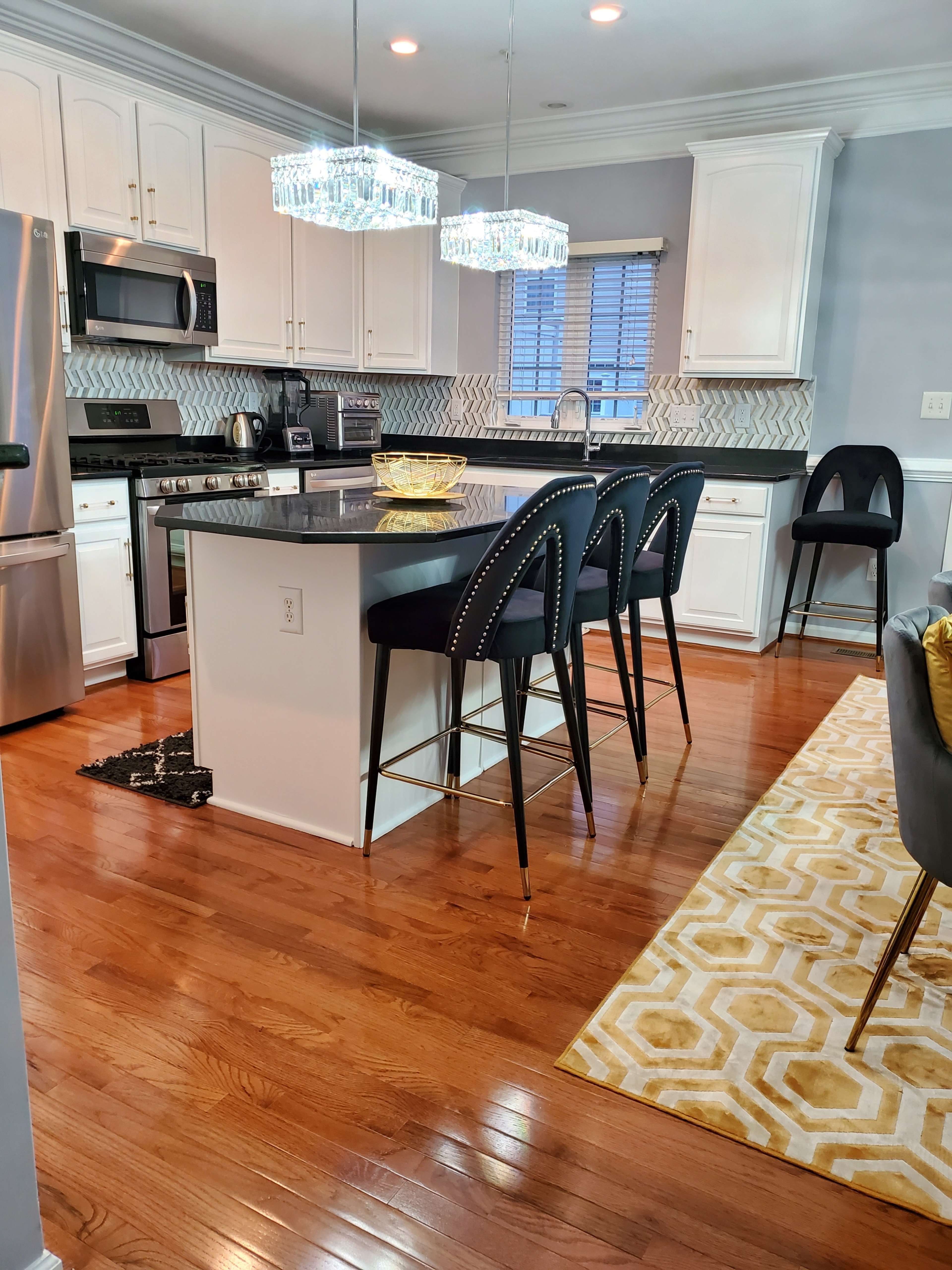 A modern kitchen with white cabinetry, stainless steel appliances, a black island with four high-backed bar stools, and a patterned rug on the hardwood floor.