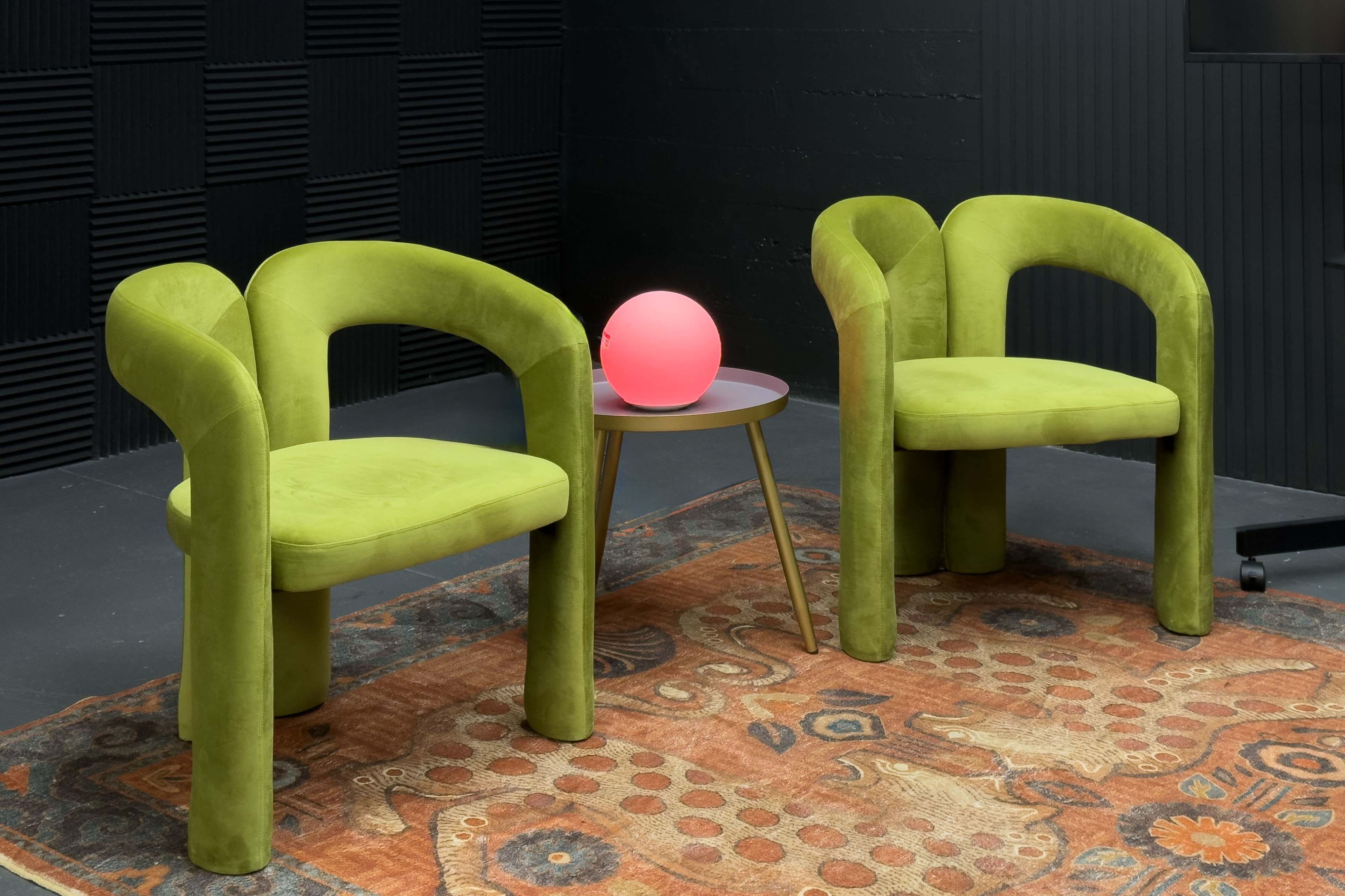 Two green upholstered chairs are positioned next to a round table with a pink orb lamp on a patterned rug.