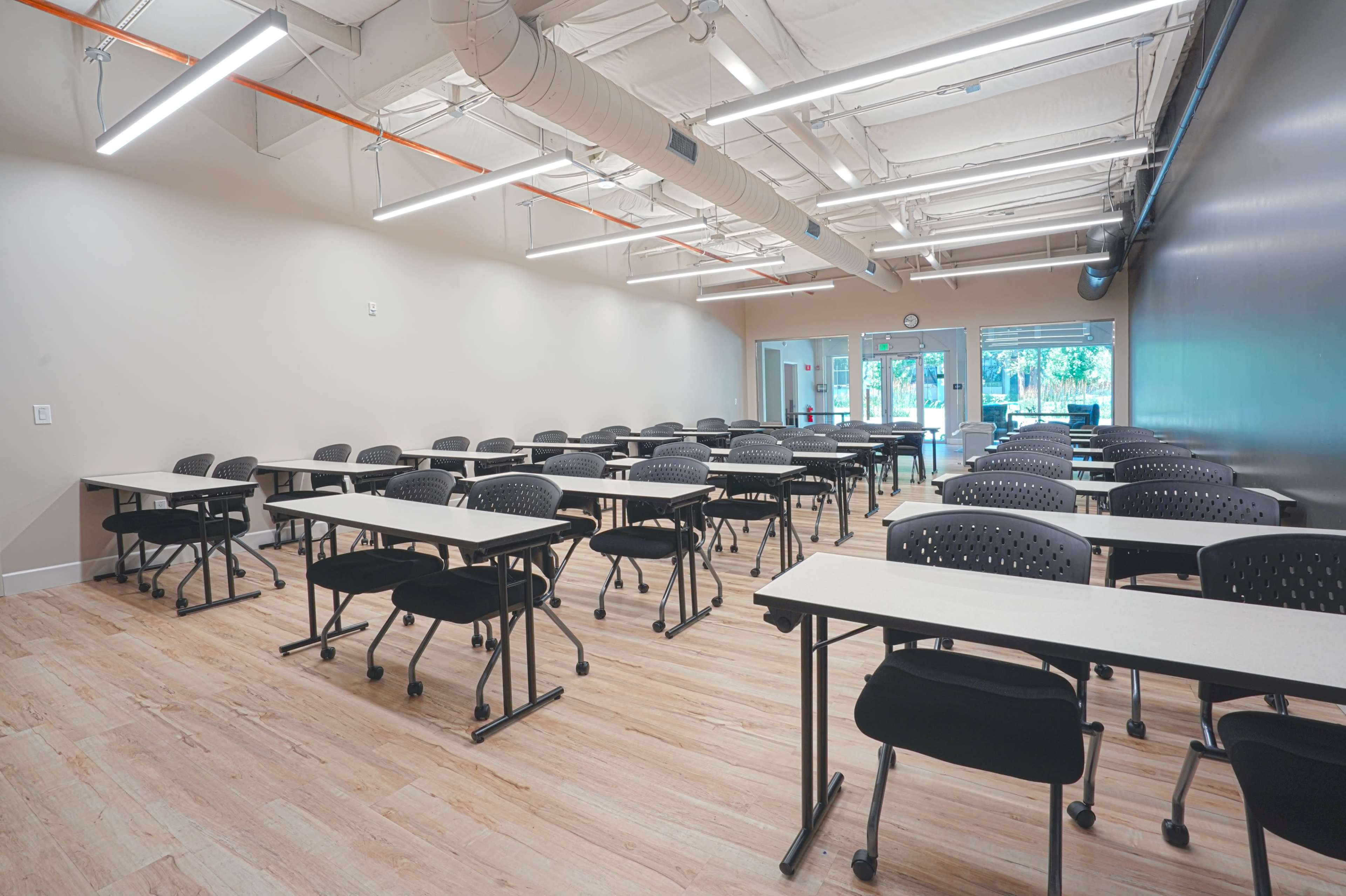A classroom with several rows of black chairs and tables arranged in a spacious layout, illuminated by overhead lights.
