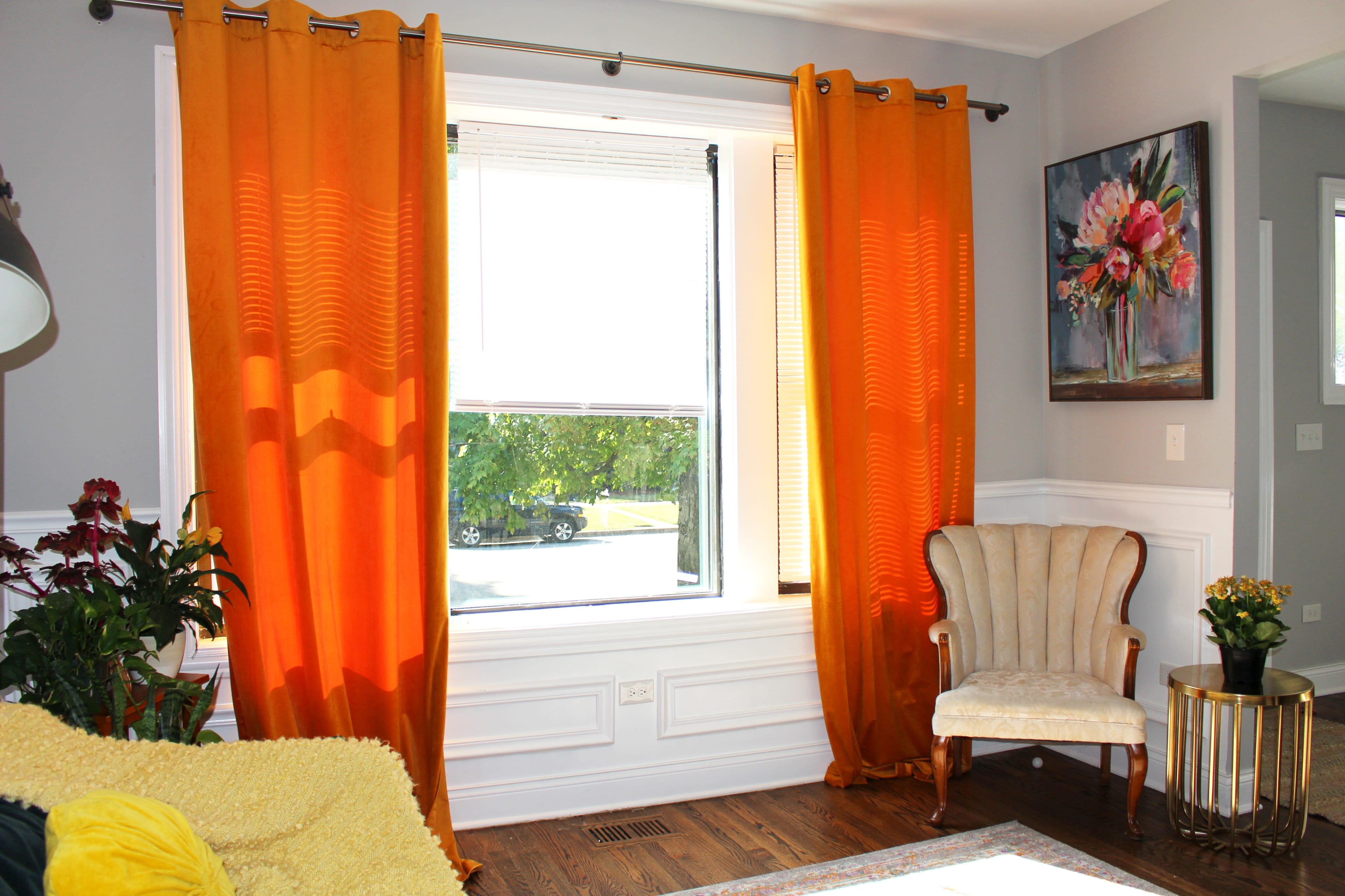 The image shows a cozy living room with orange curtains framing a window, a floral painting on the wall, a small armchair, and plants nearby.