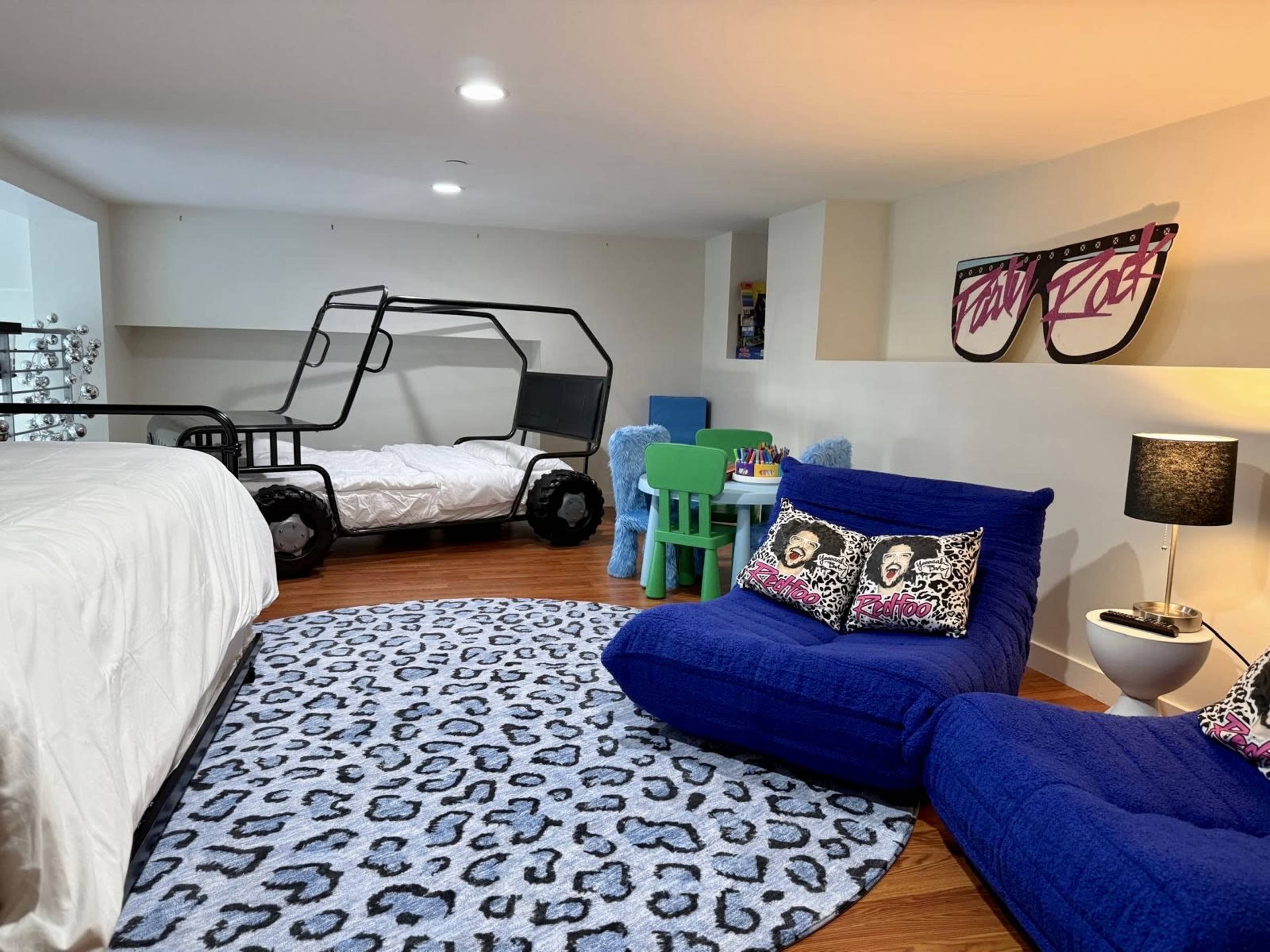 The image shows a cozy children's bedroom featuring two beds, a colorful rug, a small table with chairs, and playful decor elements.