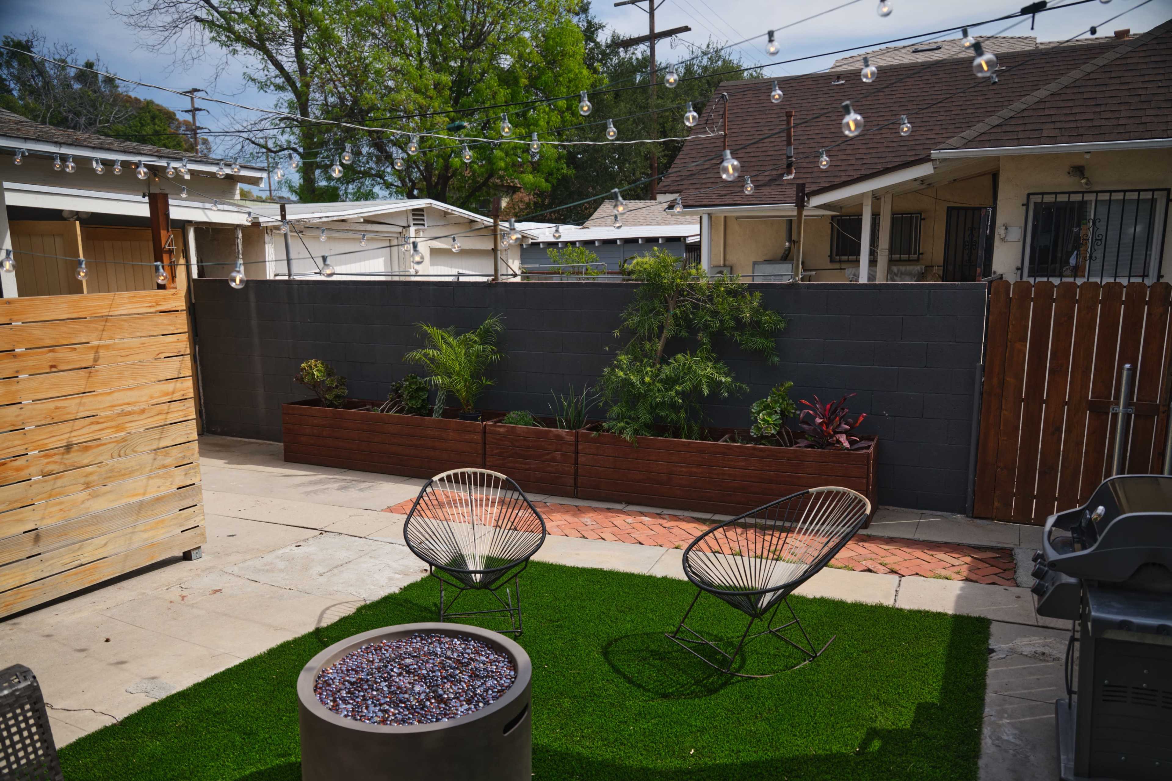 The image shows a backyard with two lounge chairs, a fire pit with decorative glass stones, and planters containing various plants, all surrounded by a wooden fence and string lights.