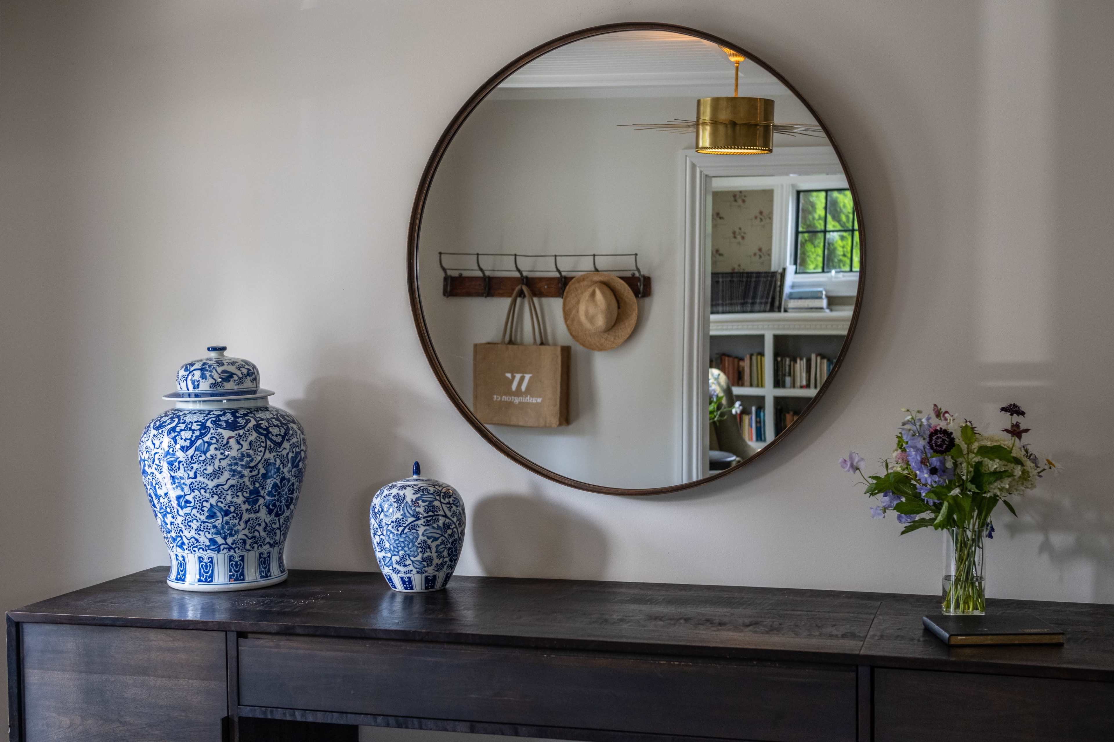 A round mirror is mounted on a wall above a dark wooden console table, flanked by blue and white porcelain vases and a bouquet of flowers in a glass vase.