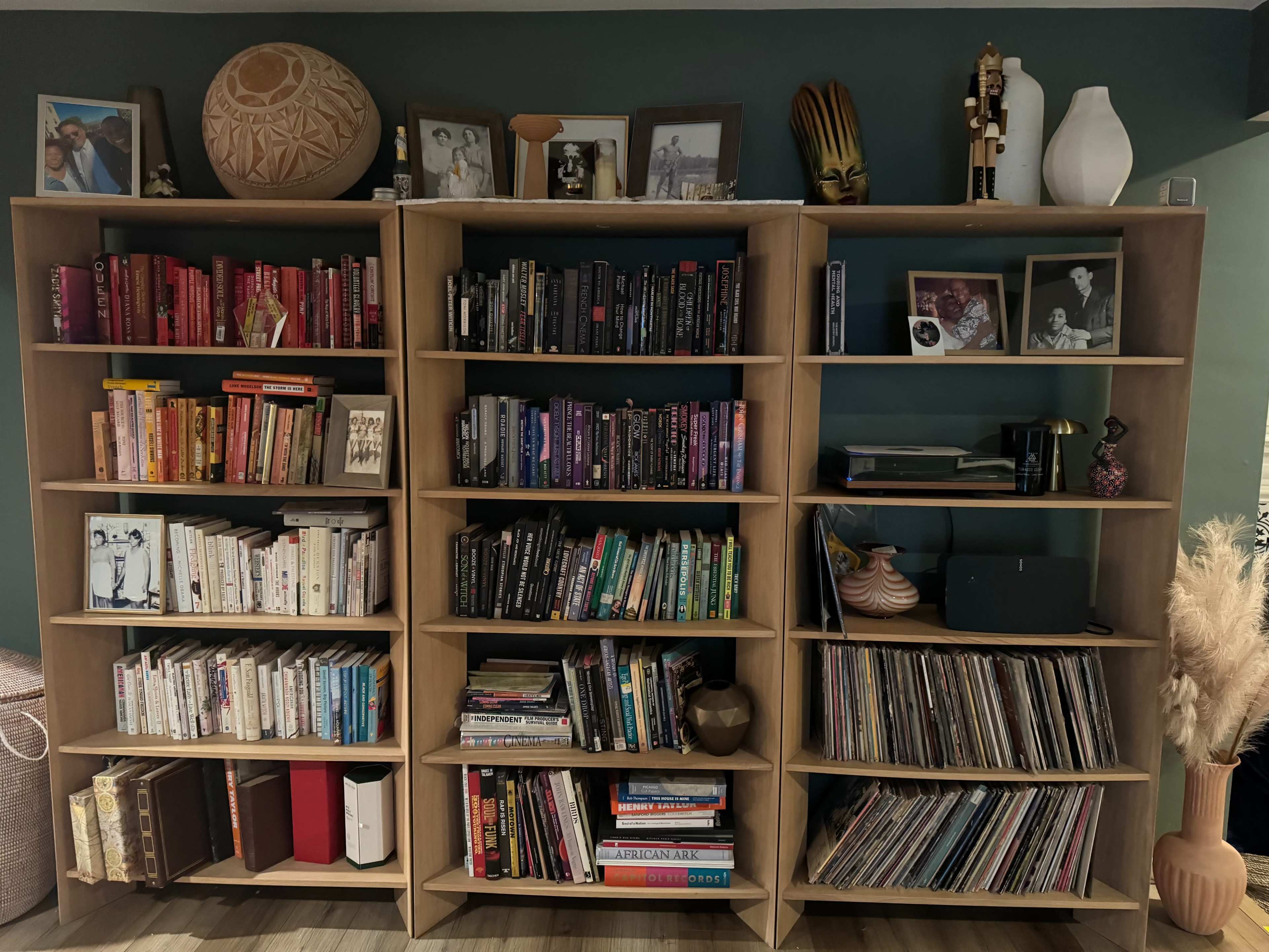 A large wooden bookshelf contains an array of books, vinyl records, and decorative items against a dark green wall.