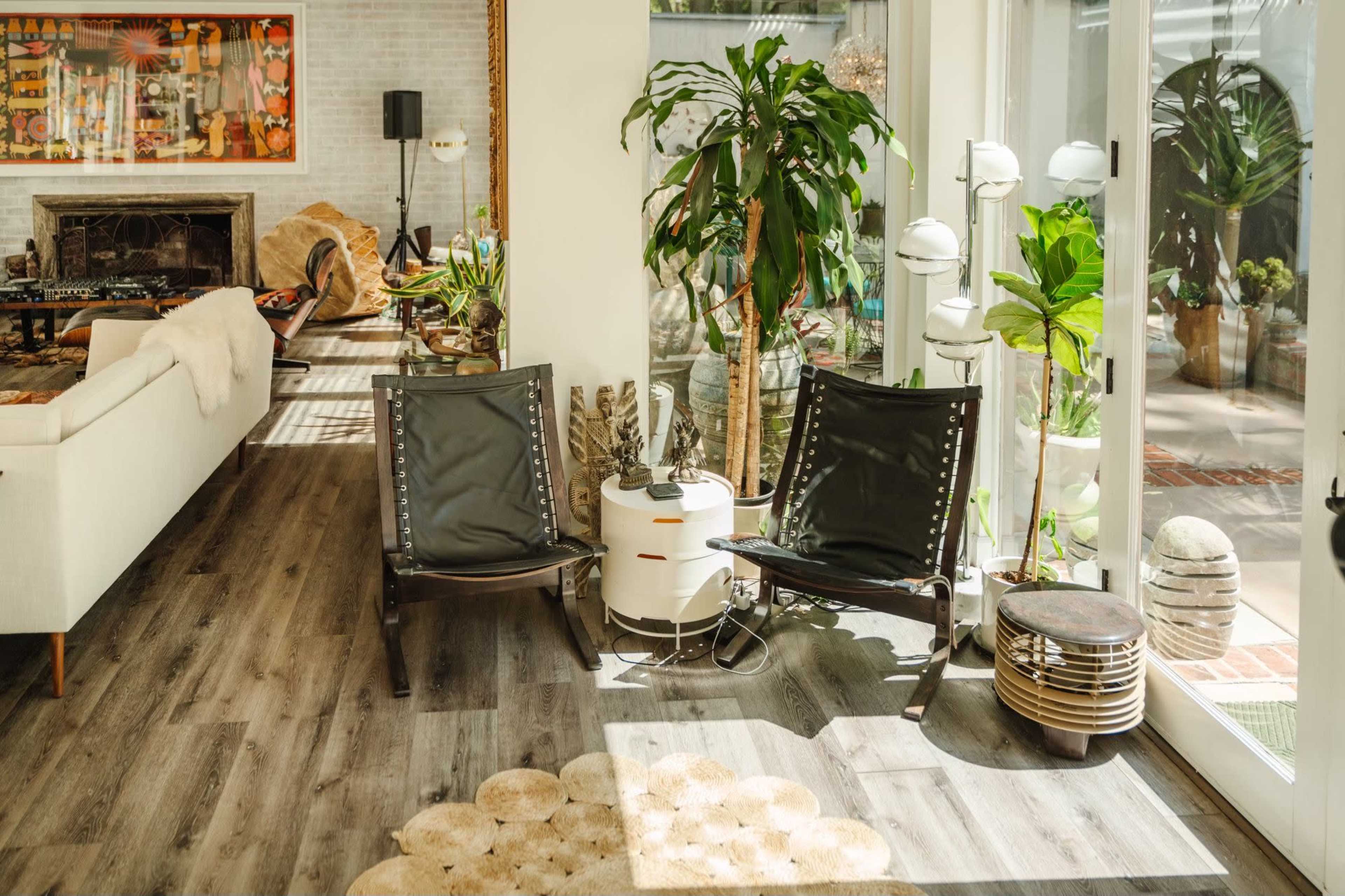The image shows a bright living space featuring two black leather chairs, a small round white table, and various plants beside large windows.