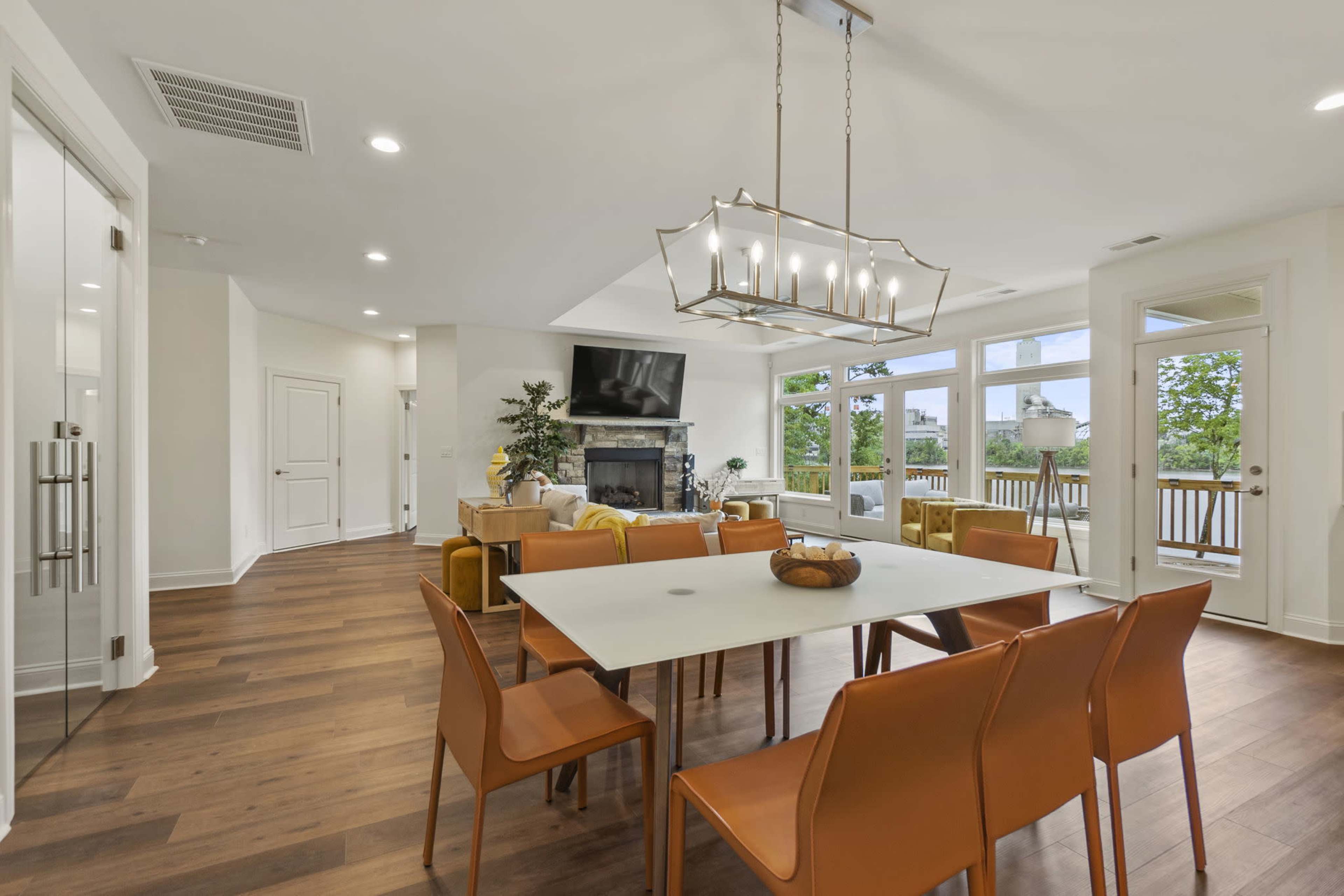 The image shows a modern dining area with a white table surrounded by orange chairs, opening up to a living room with large windows and a fireplace.