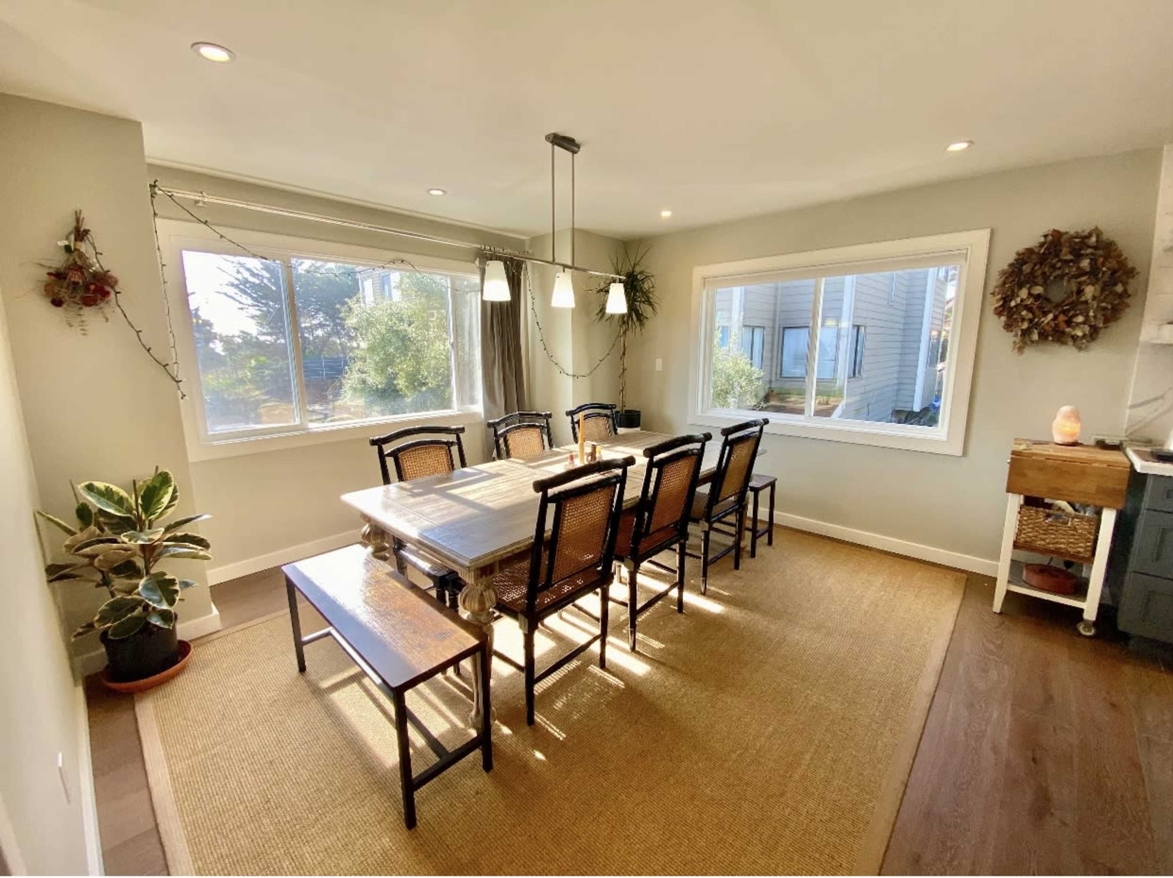 A dining area with a large wooden table surrounded by chairs, illuminated by natural light from large windows.