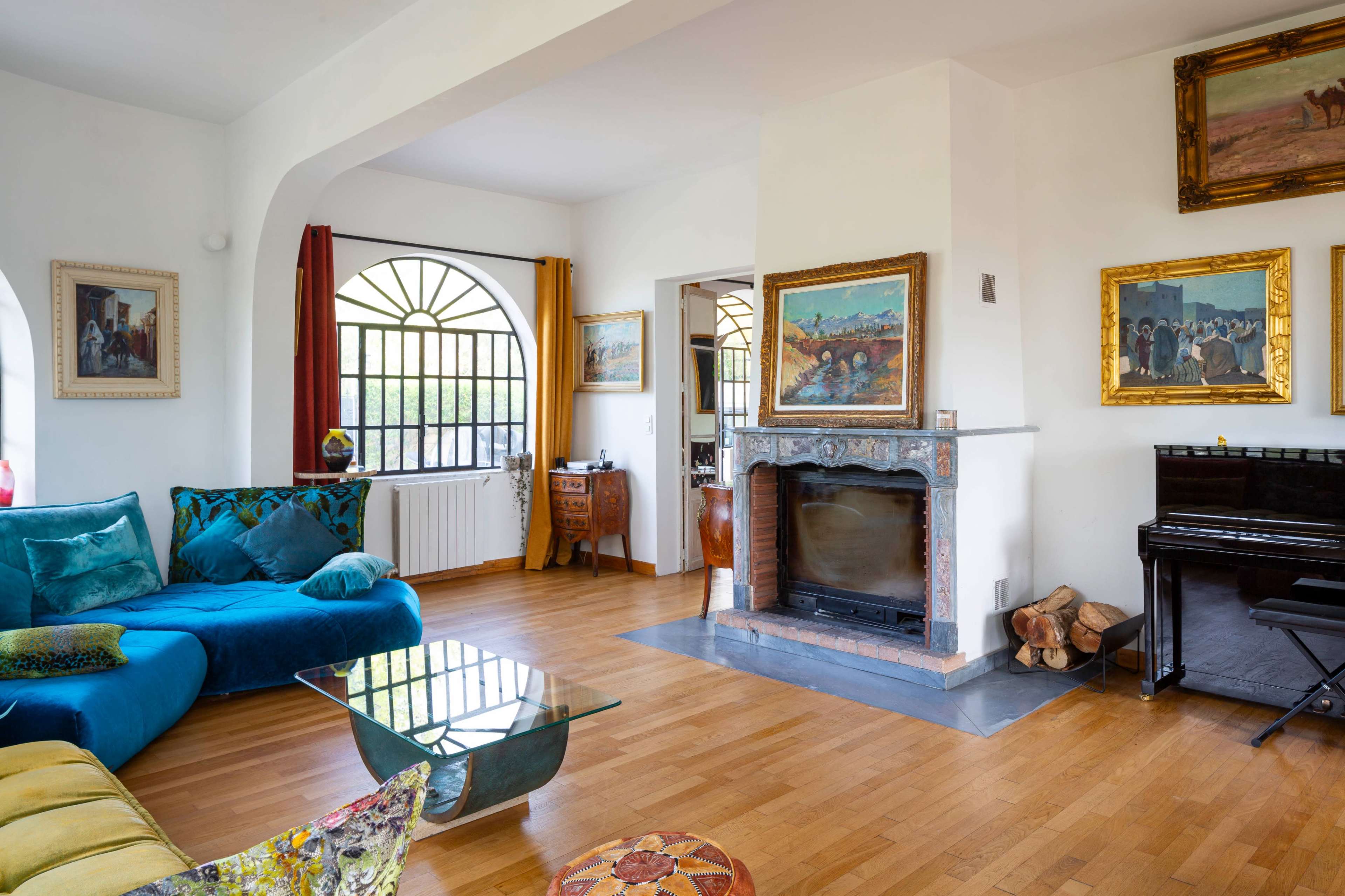 A brightly lit living room features a blue sectional sofa, a glass coffee table, a fireplace with artwork above it, and a piano against the wall adorned with various paintings.