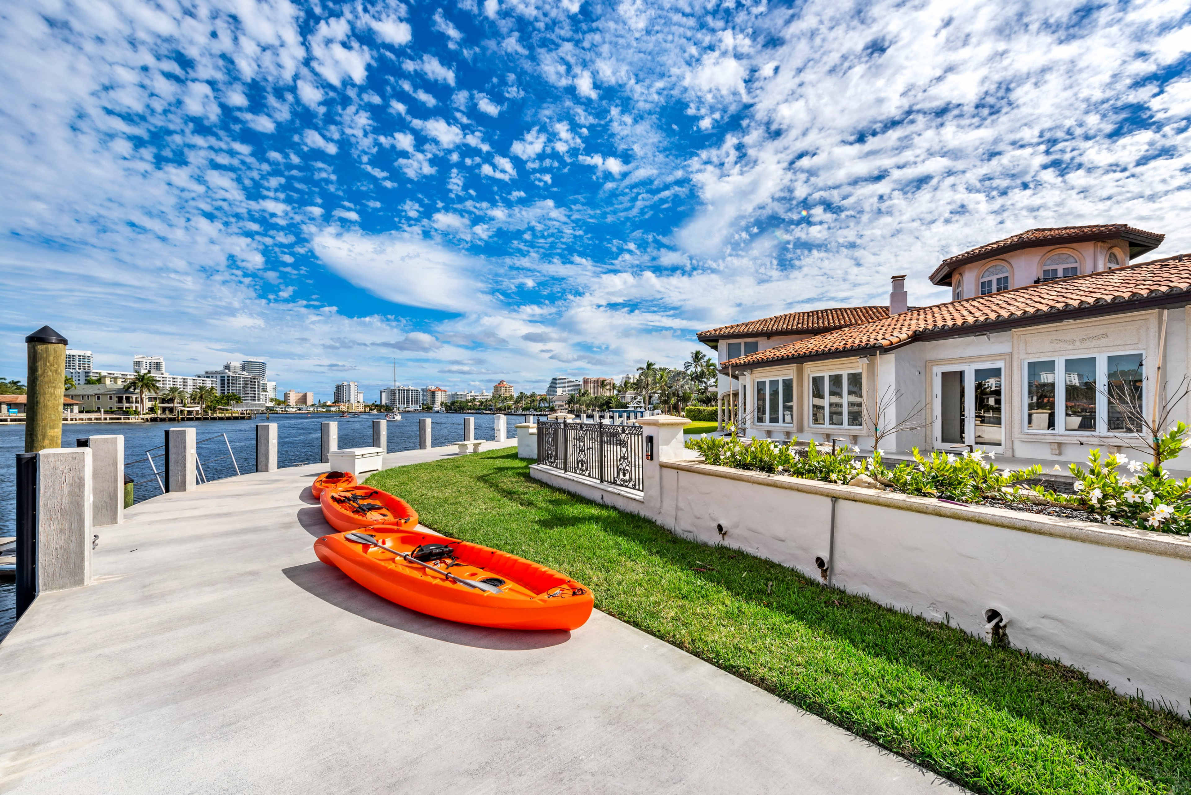 Two orange kayaks are parked on a concrete dock beside a waterfront home, with a clear sky and city skyline in the background.
