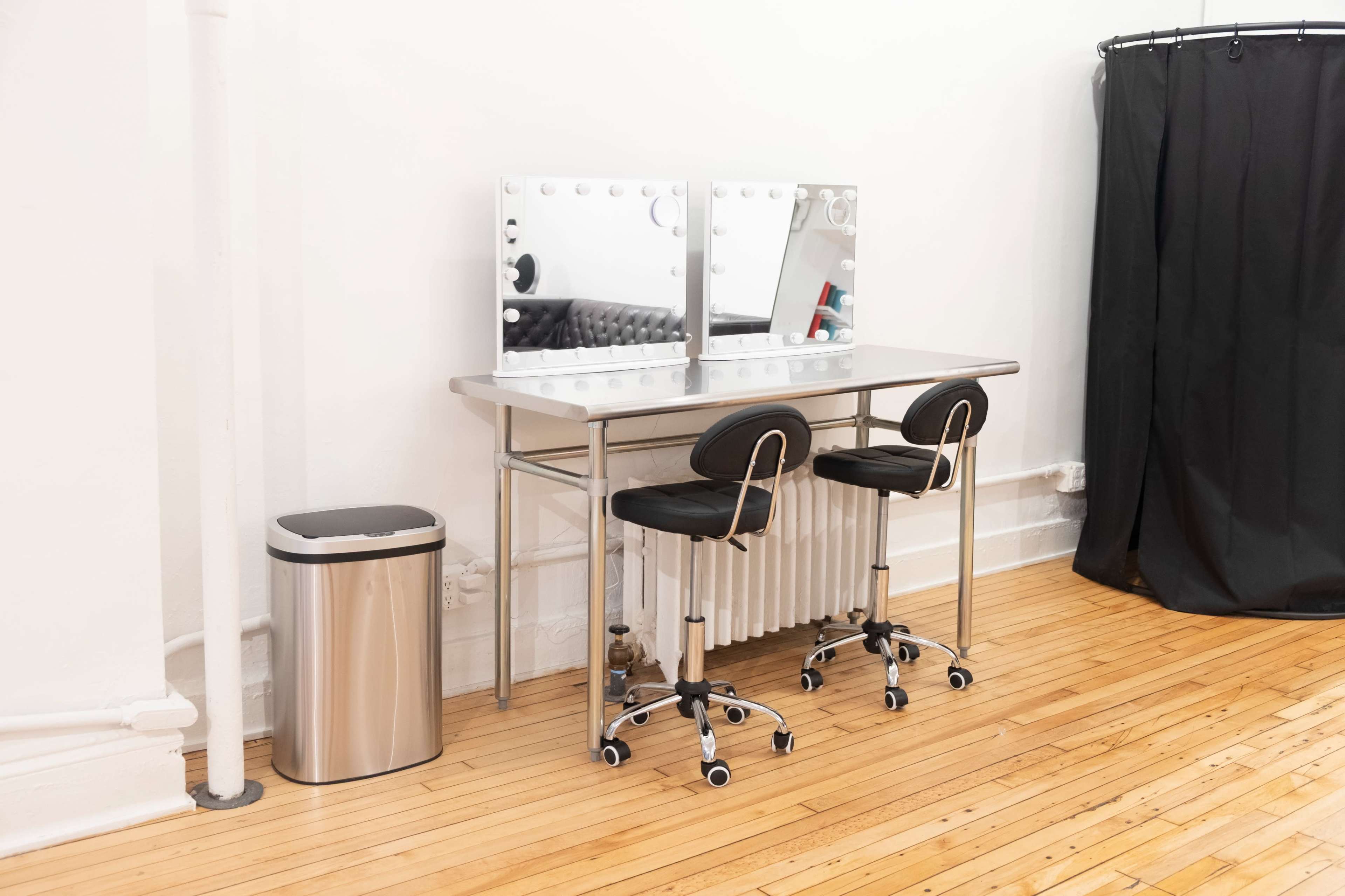 A modern makeup vanity with two rolling stools and large mirrors is positioned against a white wall, next to a metal trash can and a black curtain.