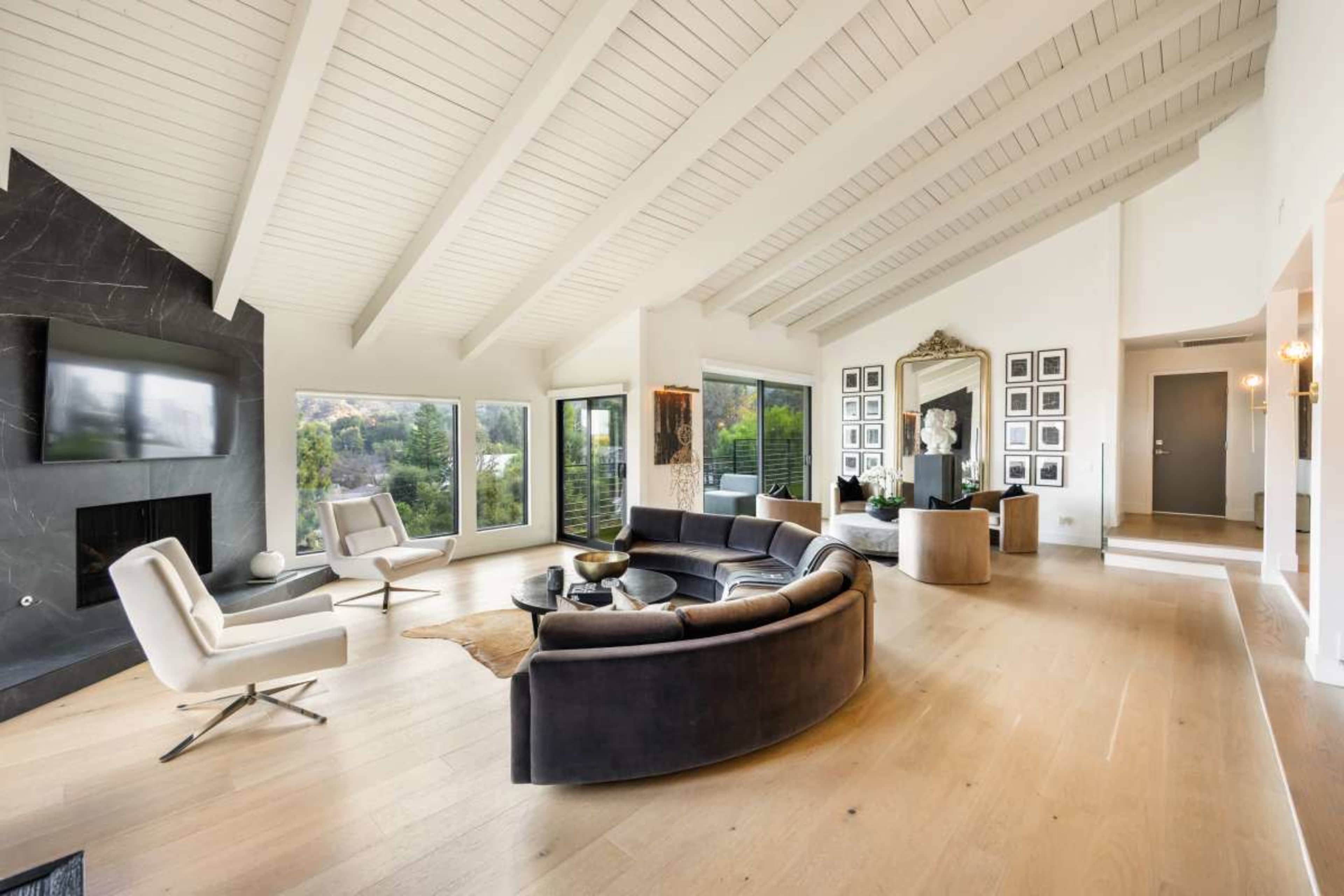 The image shows a modern living room with a curved sofa, lounge chairs, and a large mirror, featuring large windows that provide a view of the outdoors.