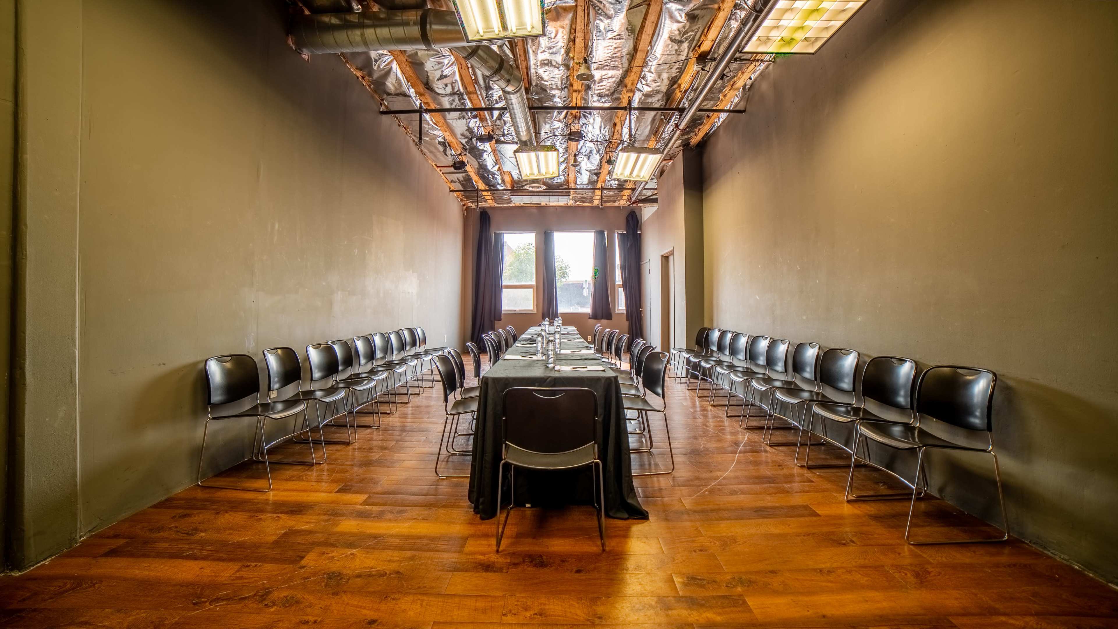 A long rectangular table is set in a narrow room with gray walls, lined with black chairs on either side and wooden flooring.