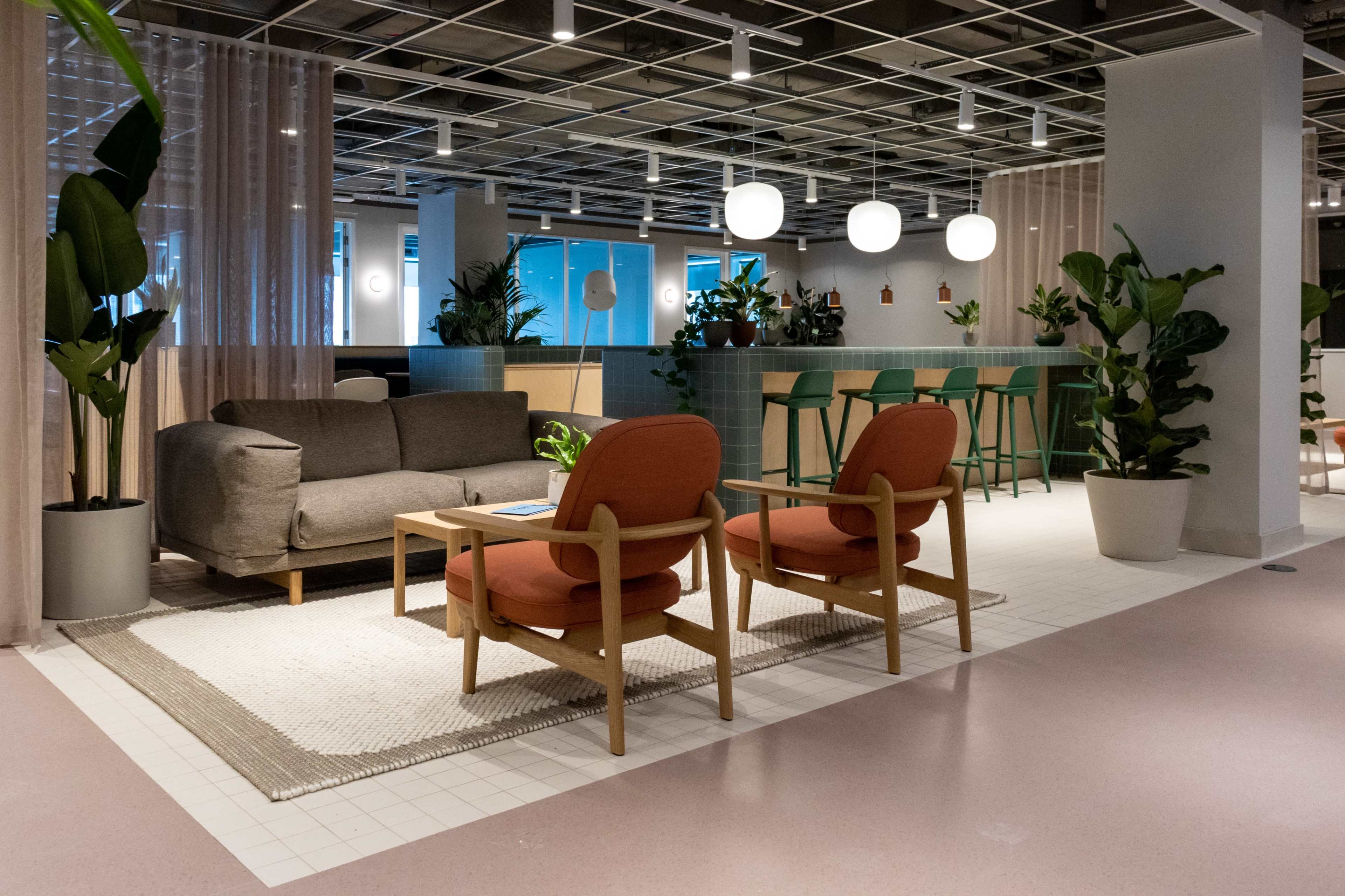 A modern lounge area featuring a gray sofa, two orange chairs, a coffee table, and a bar with green stools, surrounded by plants and soft lighting.